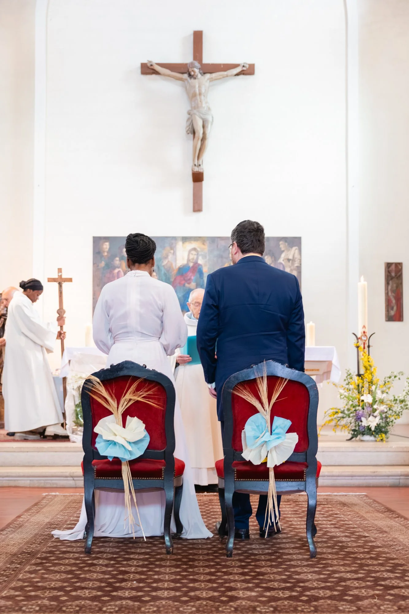 Une scène d'une cérémonie religieuse dans une église, avec deux chaises décorées de rubans et de blés, des personnes en prière et une sculpture de Jésus crucifié en arrière-plan.