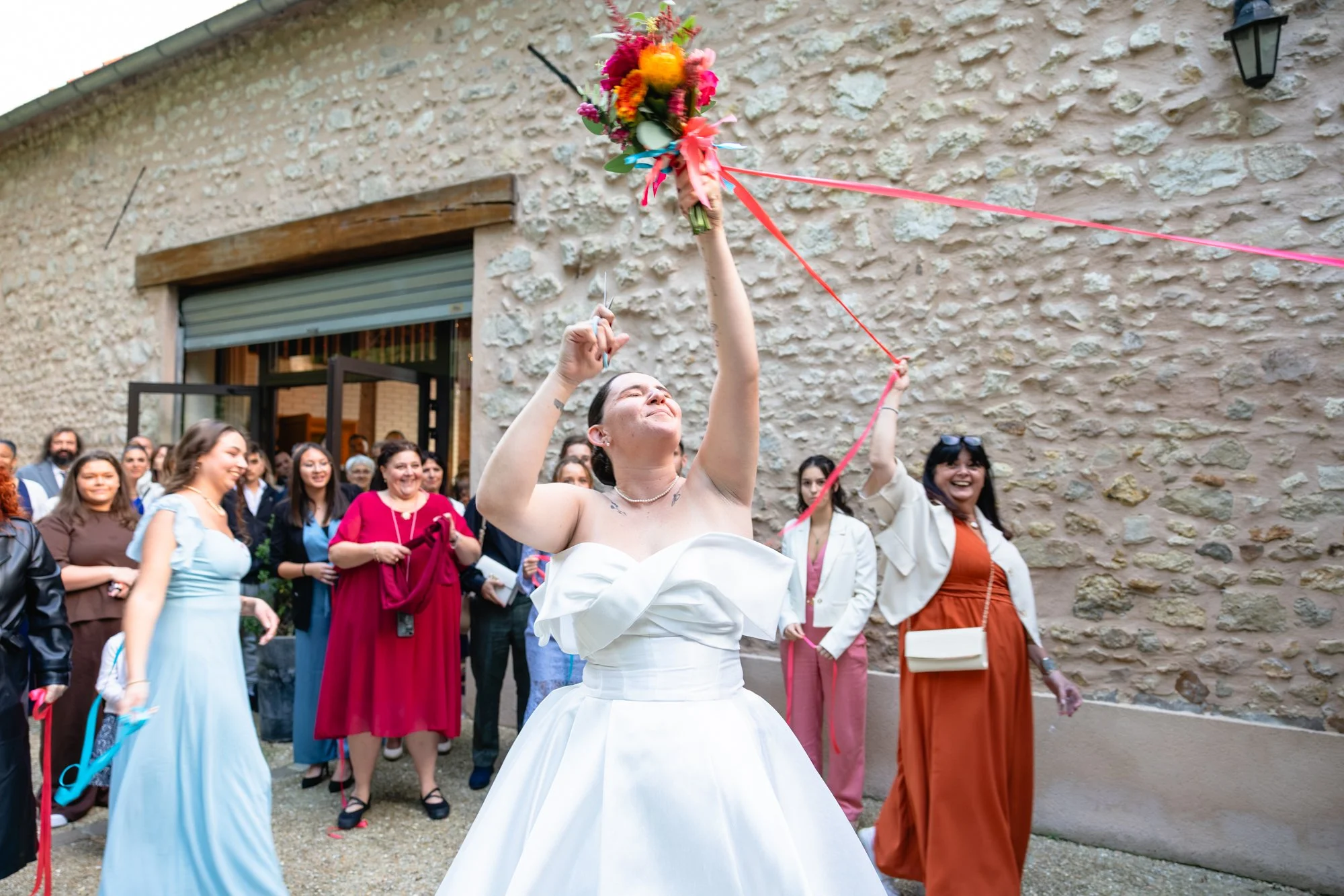 Une femme en robe blanche tient un bouquet de fleurs en l'air lors d'une célébration, entourée de plusieurs personnes souriantes, devant un mur en pierre.
