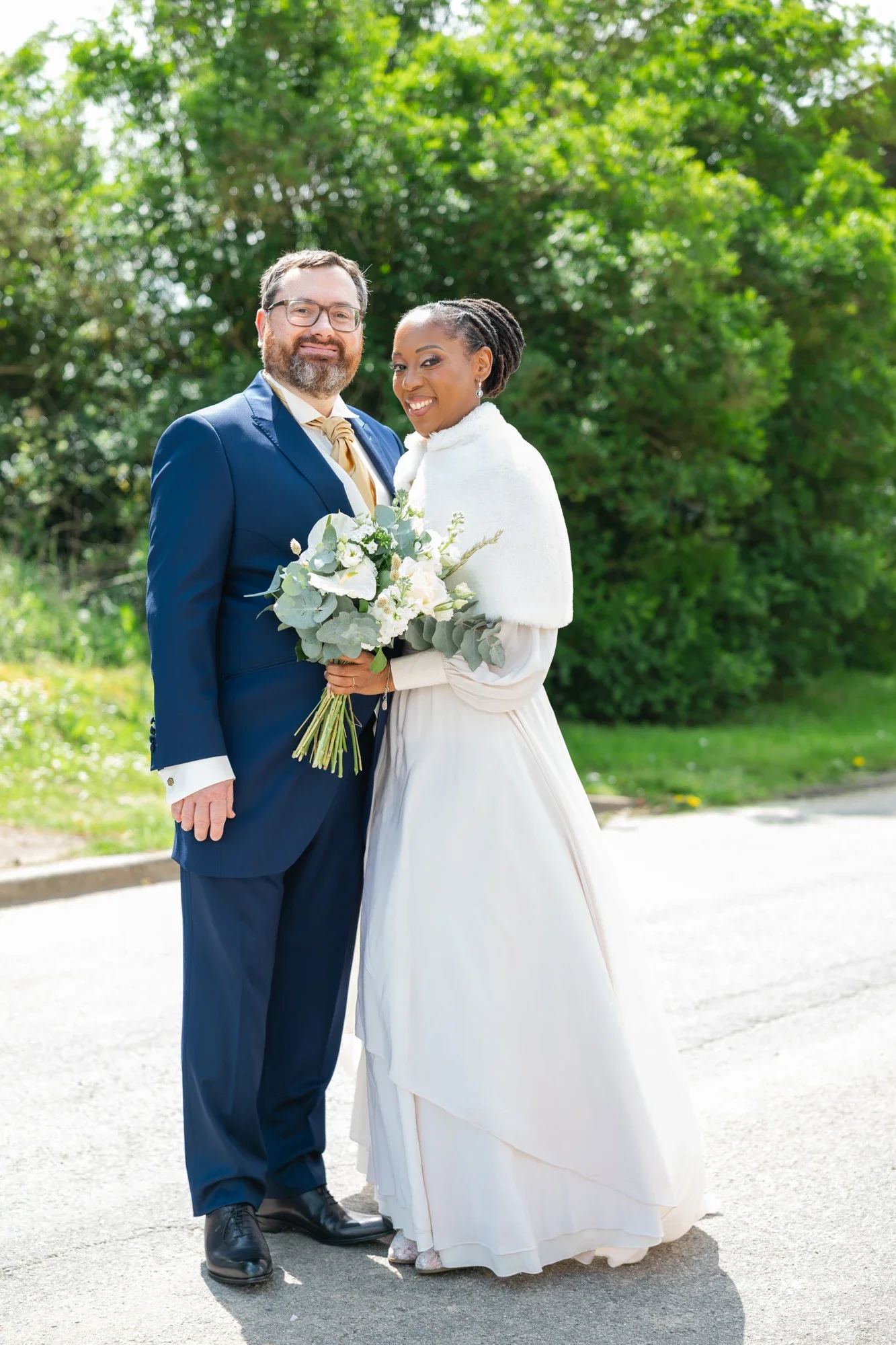 Un couple lors de leur mariage, debout sur une route bordée d'arbres verts, la femme porte une robe de mariée blanche et un manteau blanc, tenant un bouquet de fleurs. L'homme porte un costume bleu foncé avec une chemise claire et une cravate beige.