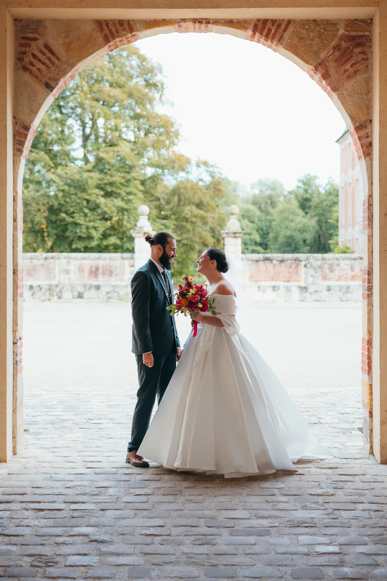 Un couple marié dans une arche en pierre, la femme porte une robe de mariée blanche avec un bouquet de fleurs colorées, et l'homme porte un costume noir avec une cravate, dans un décor extérieur avec des arbres verts en arrière-plan.