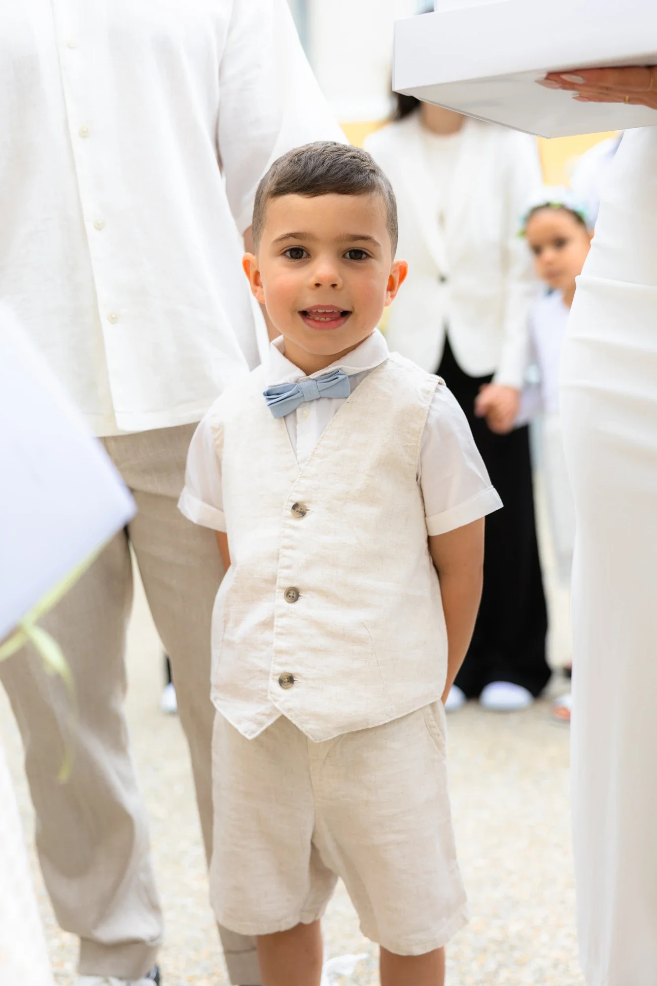 Un jeune garçon habillé en beige avec un nœud papillon gris clair, lors d'une cérémonie ou occasion formelle, entouré d'adultes également habillés en blanc.