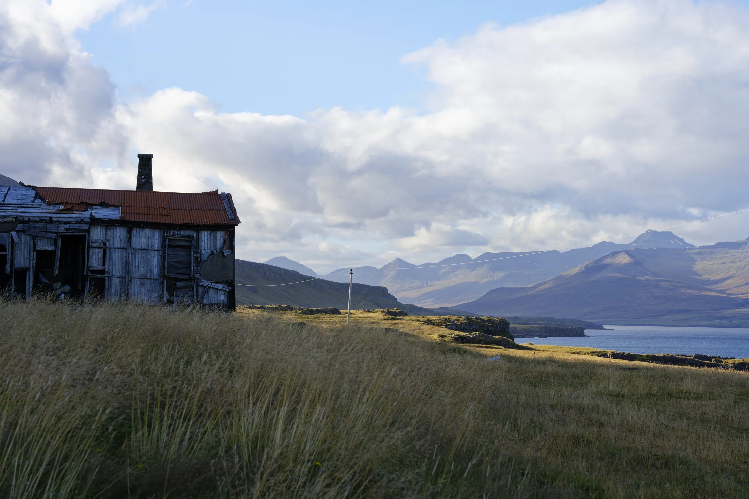 An old, weathered wooden house with a rusty tin roof situated in a grassy field next to a large body of water, with mountains in the distance under a partly cloudy sky.