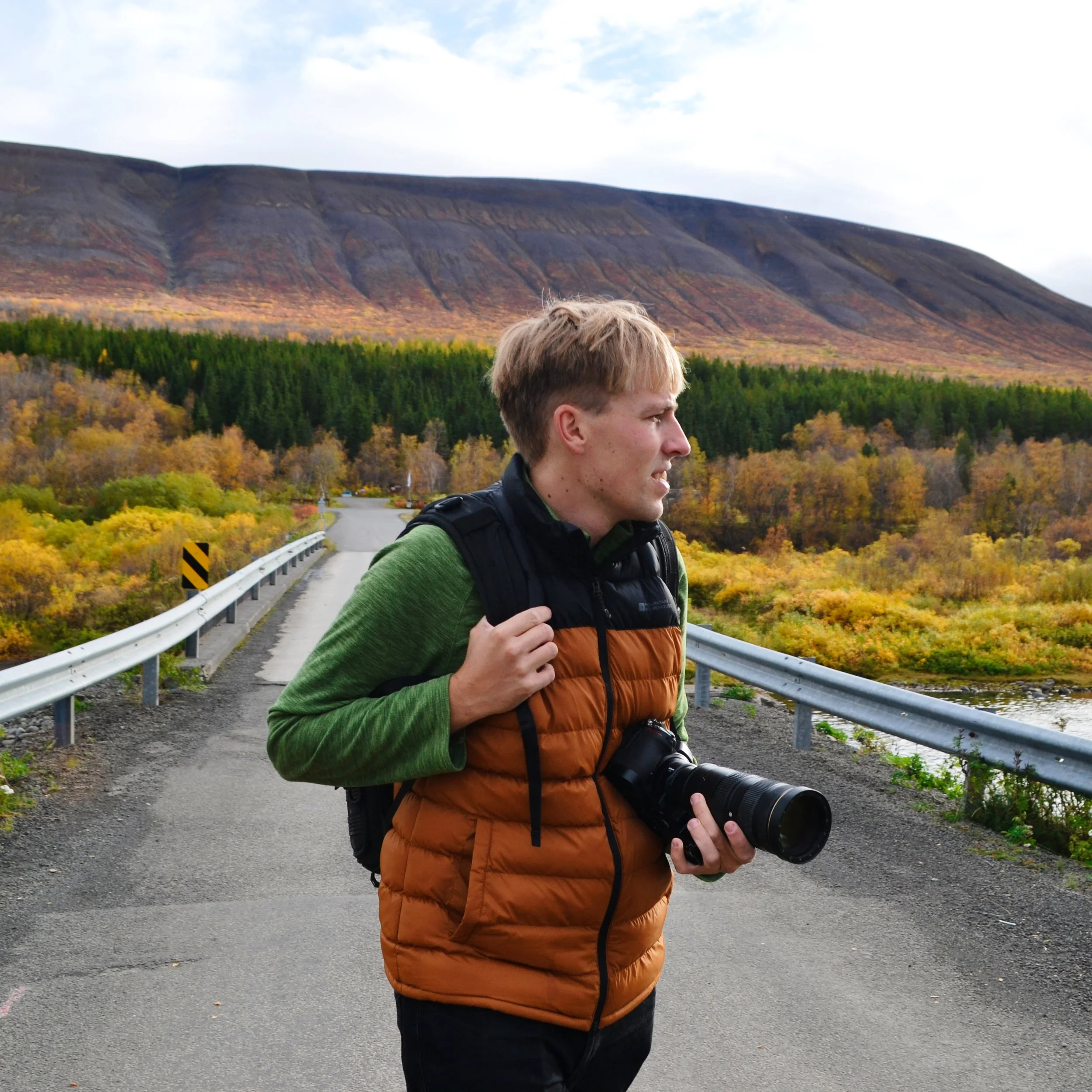 A young man with short blond hair is walking outdoors along a paved road. He is carrying a professional camera with a large lens and has a camera strap around his neck. He is wearing a green long-sleeve shirt, a brown puffer vest, and dark pants. In the background, there are colorful autumn trees and a mountain range under a partly cloudy sky.