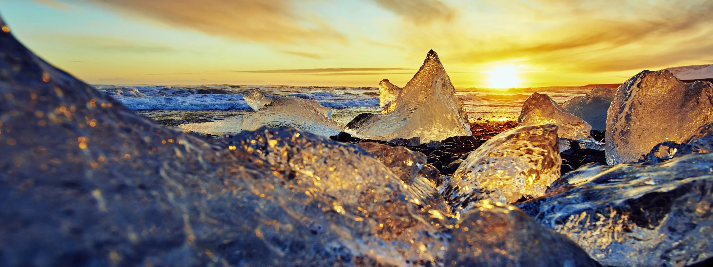 Icebergs on a beach at sunset with the sun near the horizon and a colorful sky.