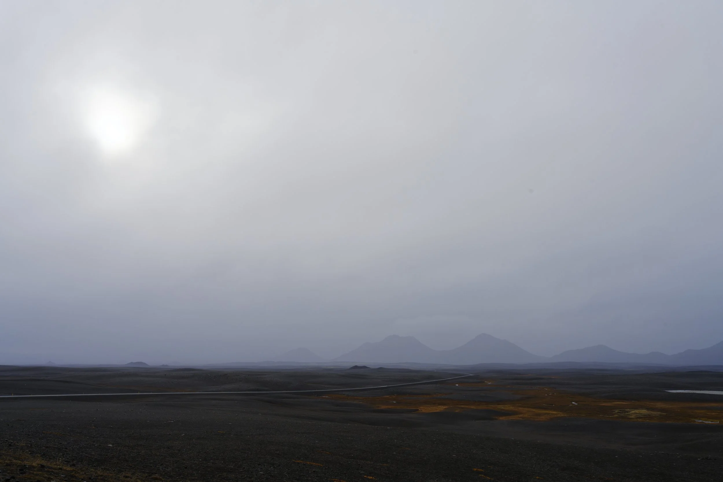 A vast barren landscape with dark soil stretching to distant mountains under a cloudy sky with the sun faintly visible.