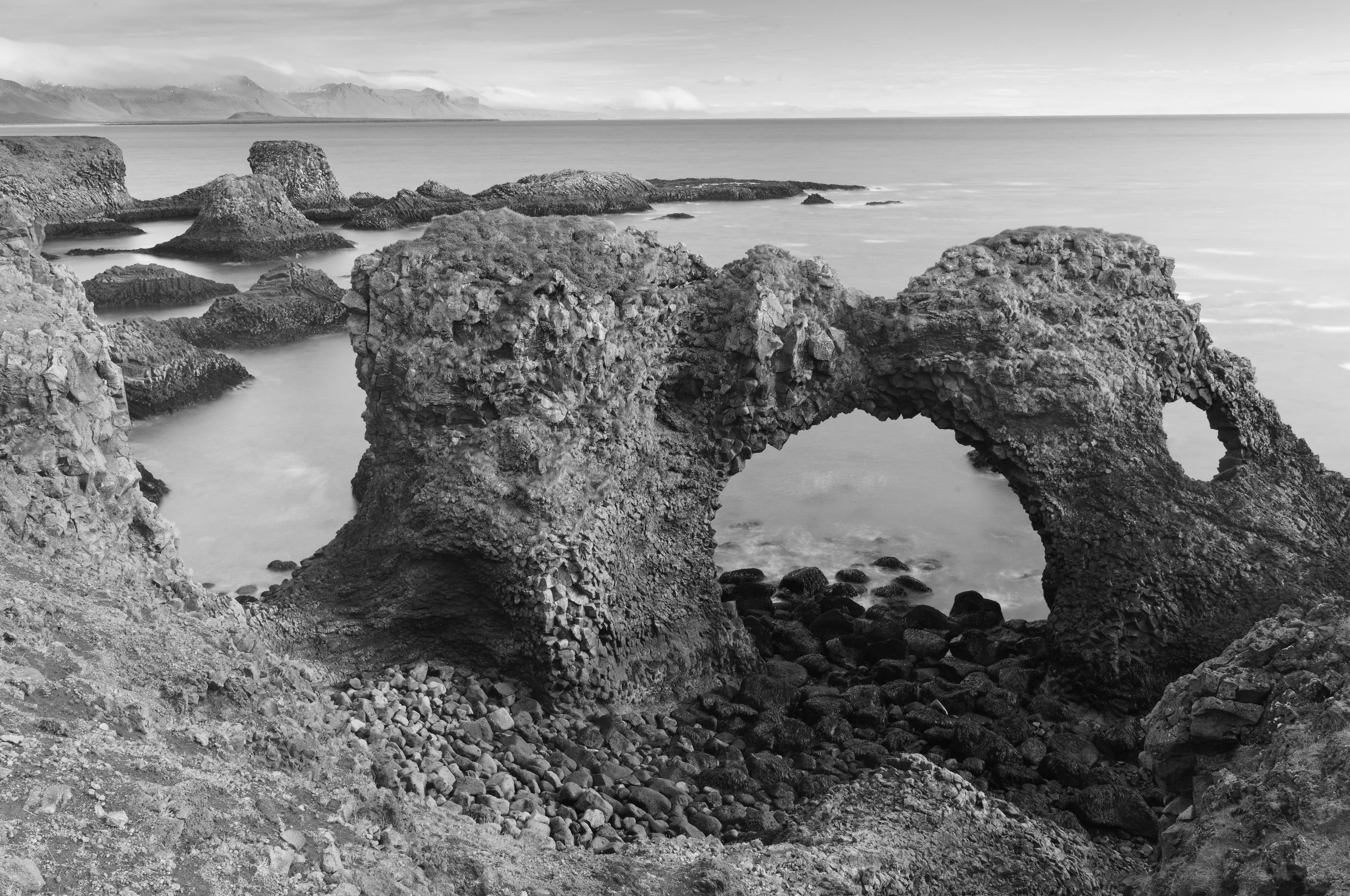 Black and white photo of rocky coastal landscape with concrete arch formation, water surrounding rocks, and distant mountains on the horizon.