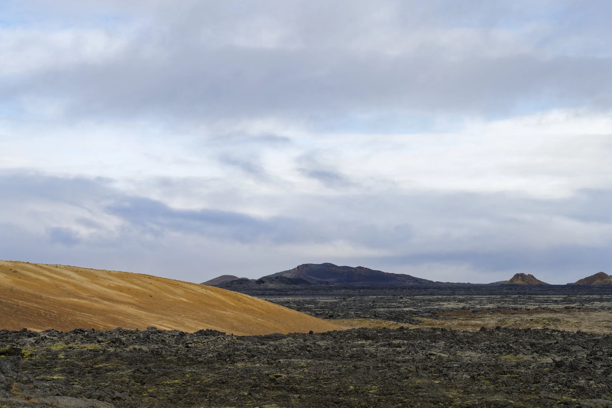 Volcanic landscape with dark, rocky foreground, rolling golden hills, distant dark mountains, and a cloudy sky.
