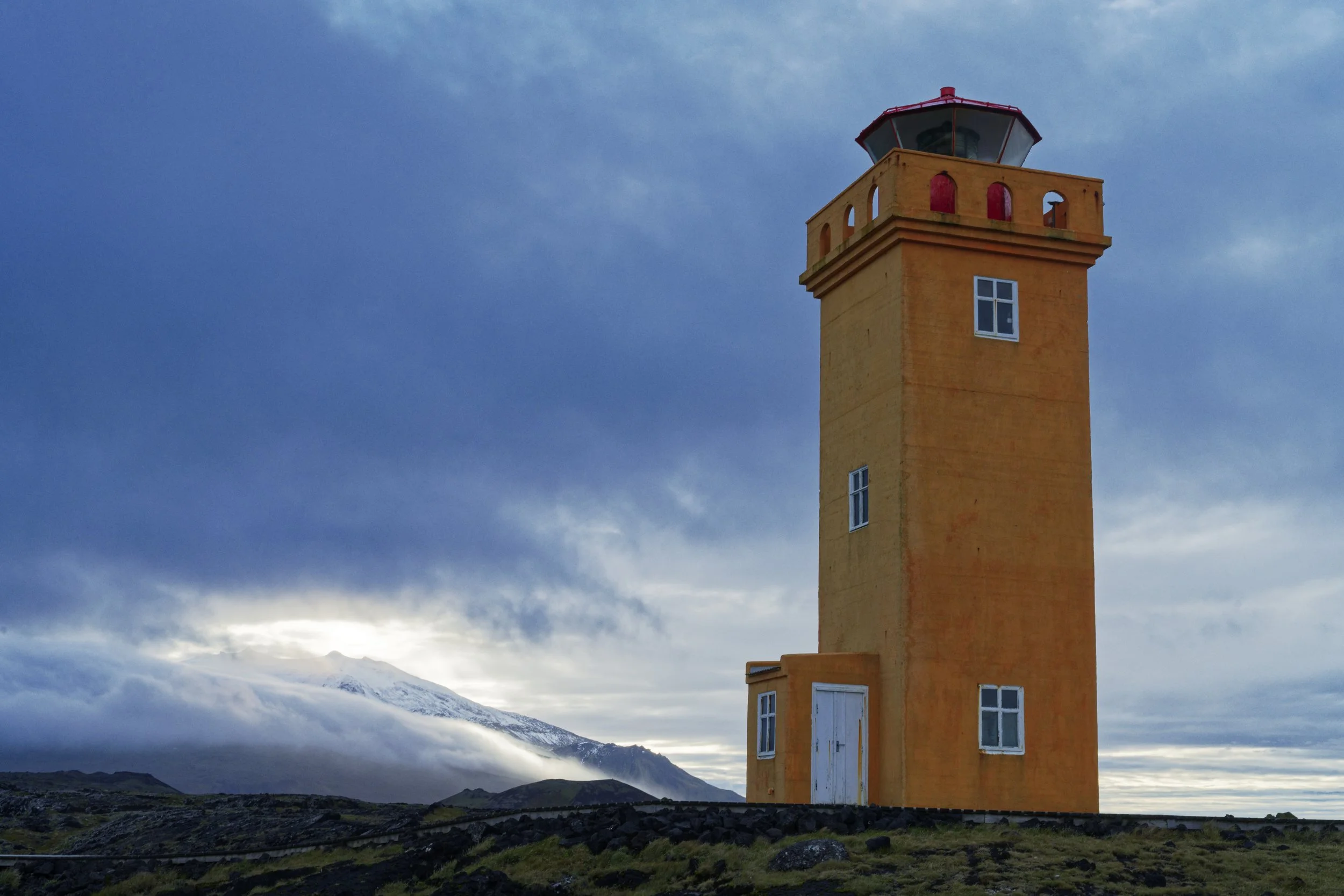 A tall, orange lighthouse with a red top stands against a cloudy sky. Snow-capped mountains are visible in the background with low clouds around their peaks.