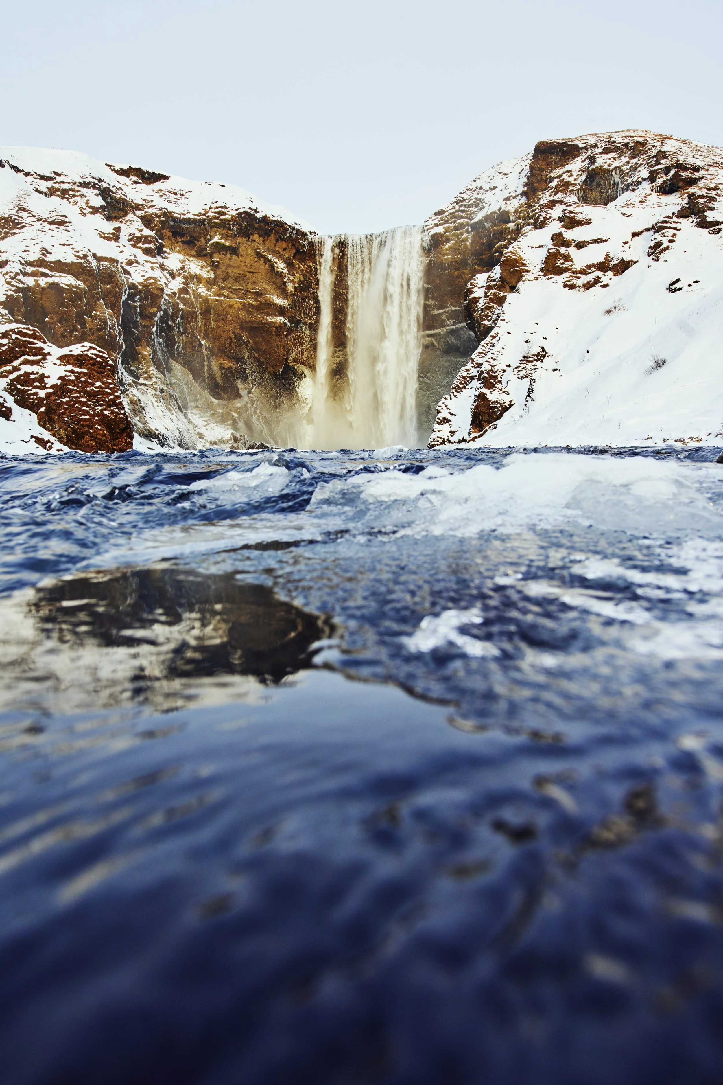 Waterfall surrounded by snow-covered rocks with a clear blue sky overhead.