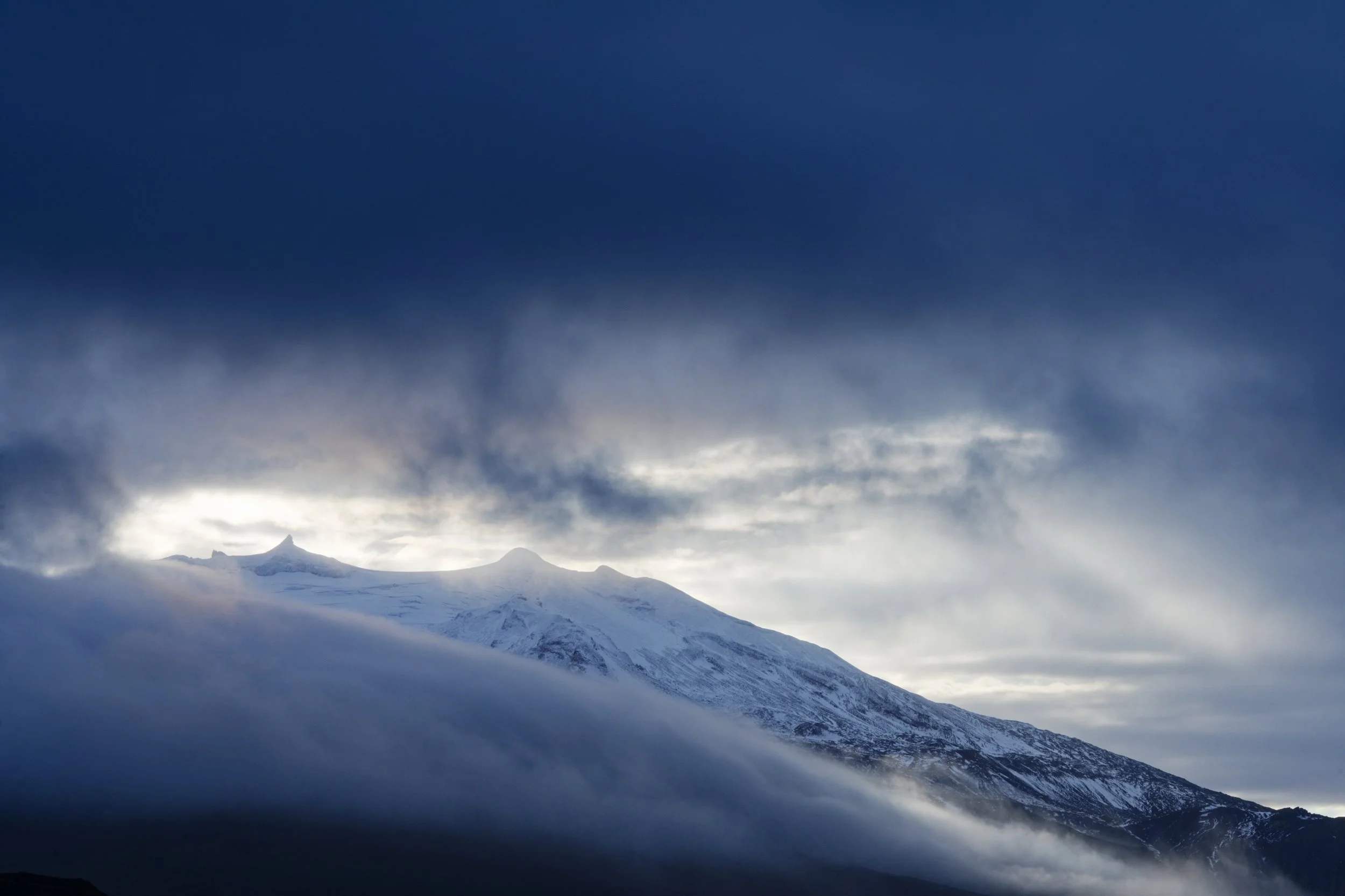 Snow-covered mountain peaks beneath a dark, cloudy sky with sunlight breaking through.