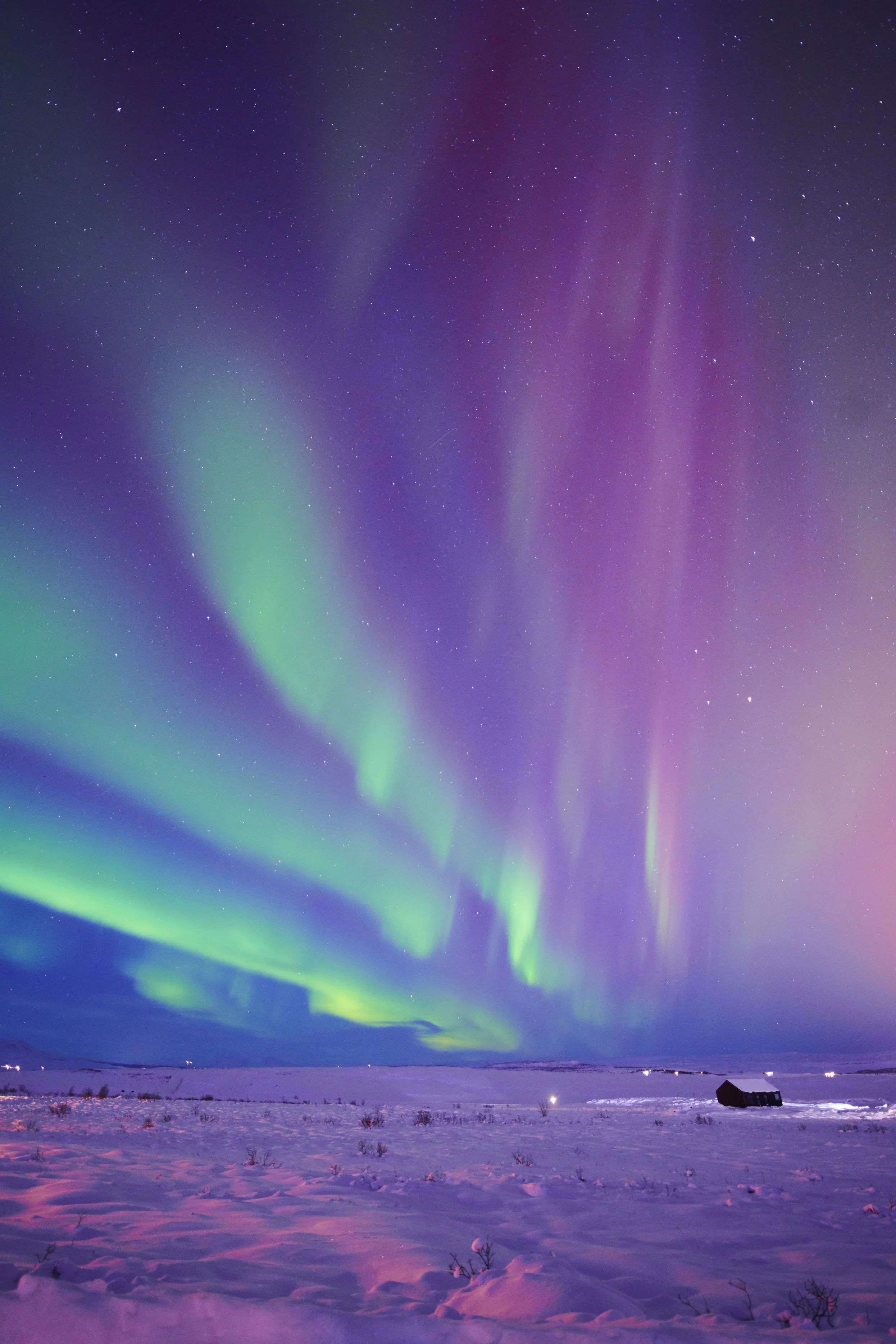 Night sky illuminated with colorful aurora borealis over snowy landscape, with a small dark house in the distance.