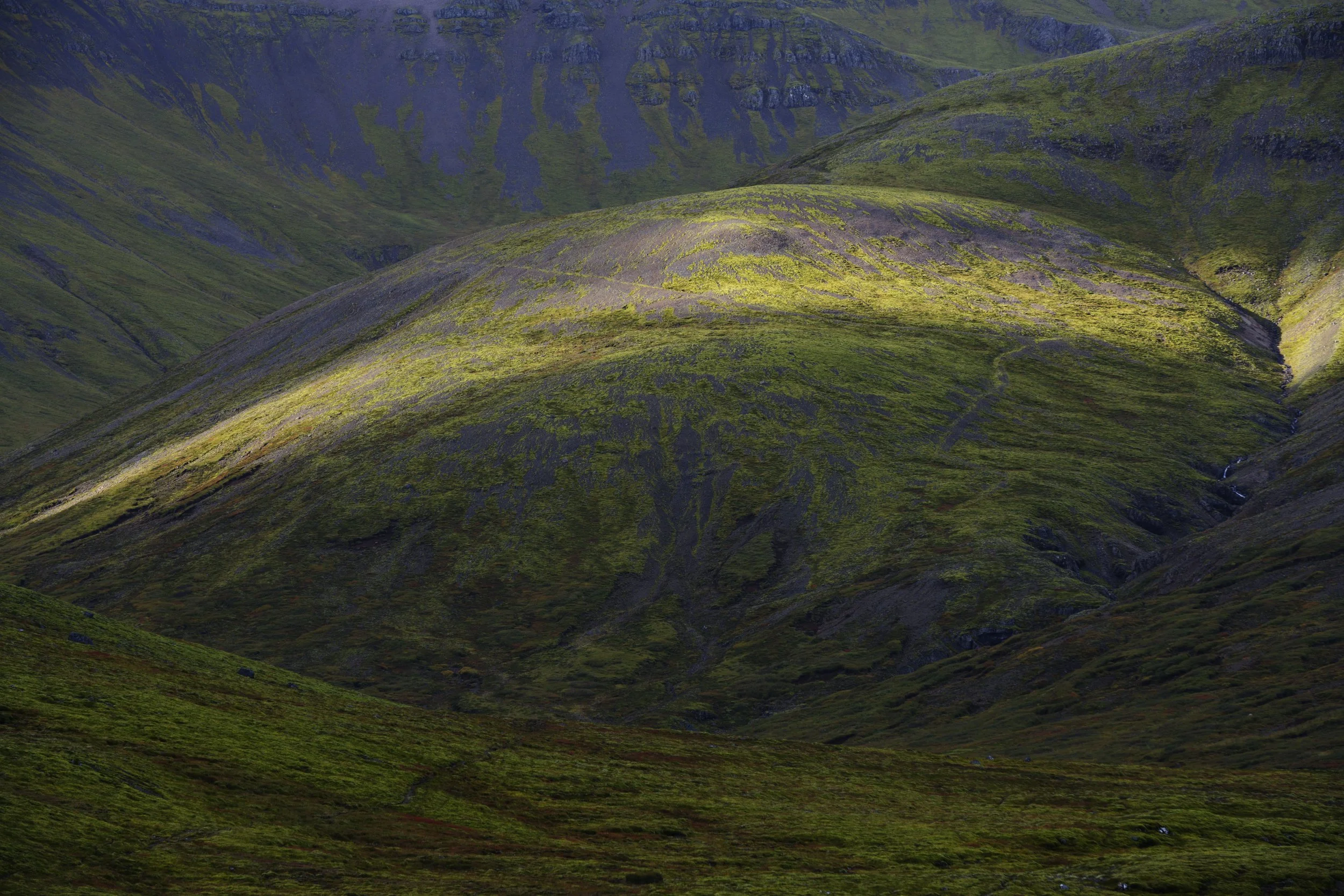 Green mountainous landscape with steep slopes, patches of moss and grass, and a small waterfall running down the hillside.