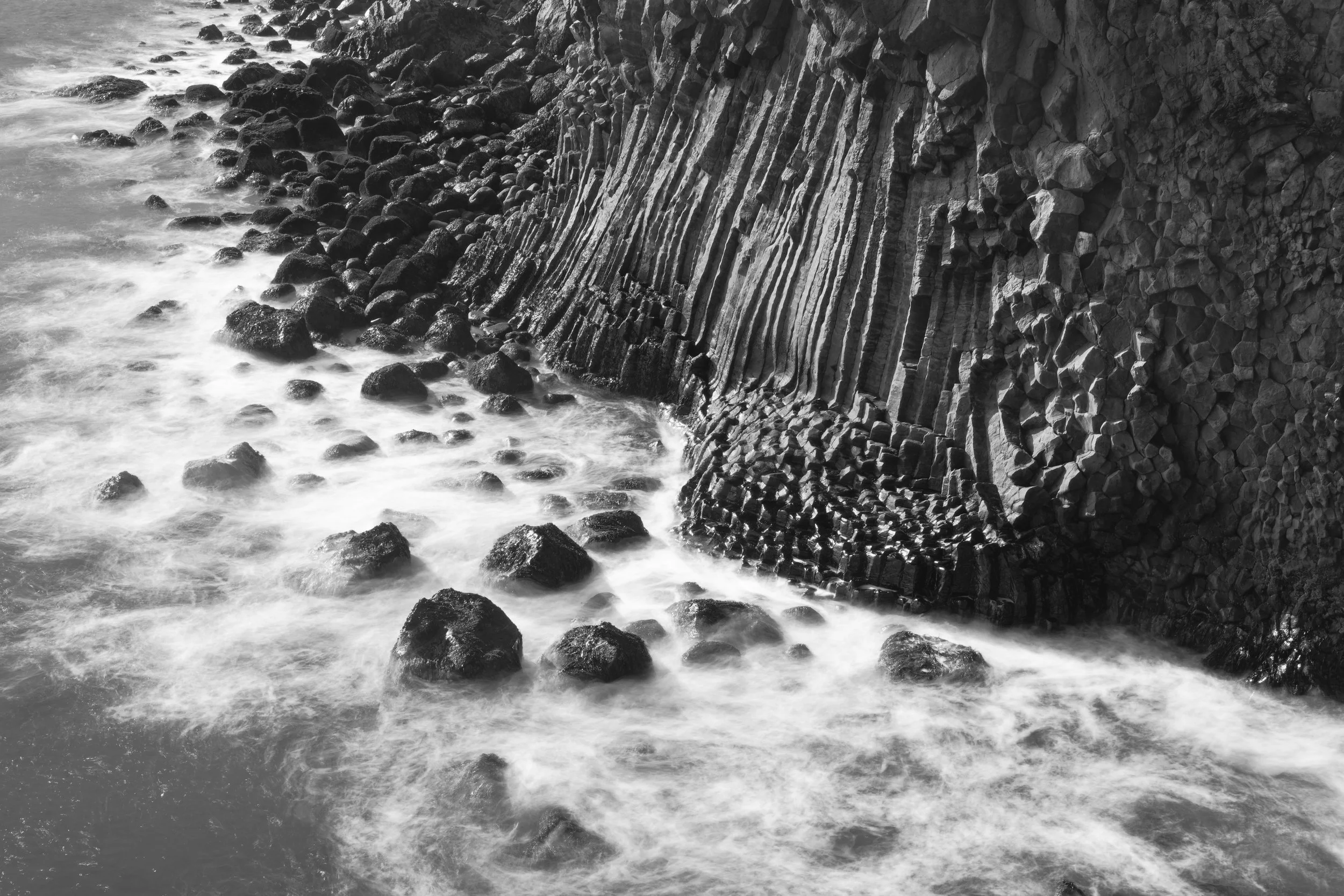 Black and white photo of a rocky shoreline with water washing over the rocks, and a cliff with layered rock formations on the right side.