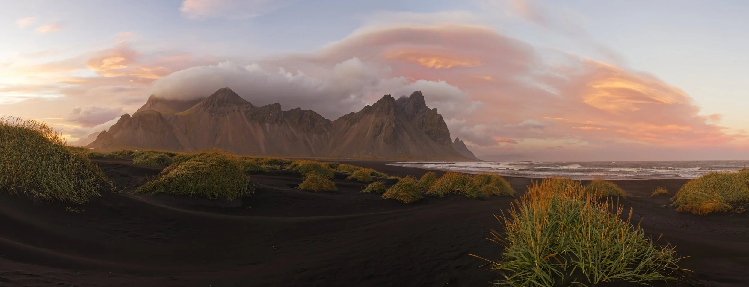 A coastal landscape with black sand dunes, green grass, and mountains in the background during sunset with colorful sky and clouds.