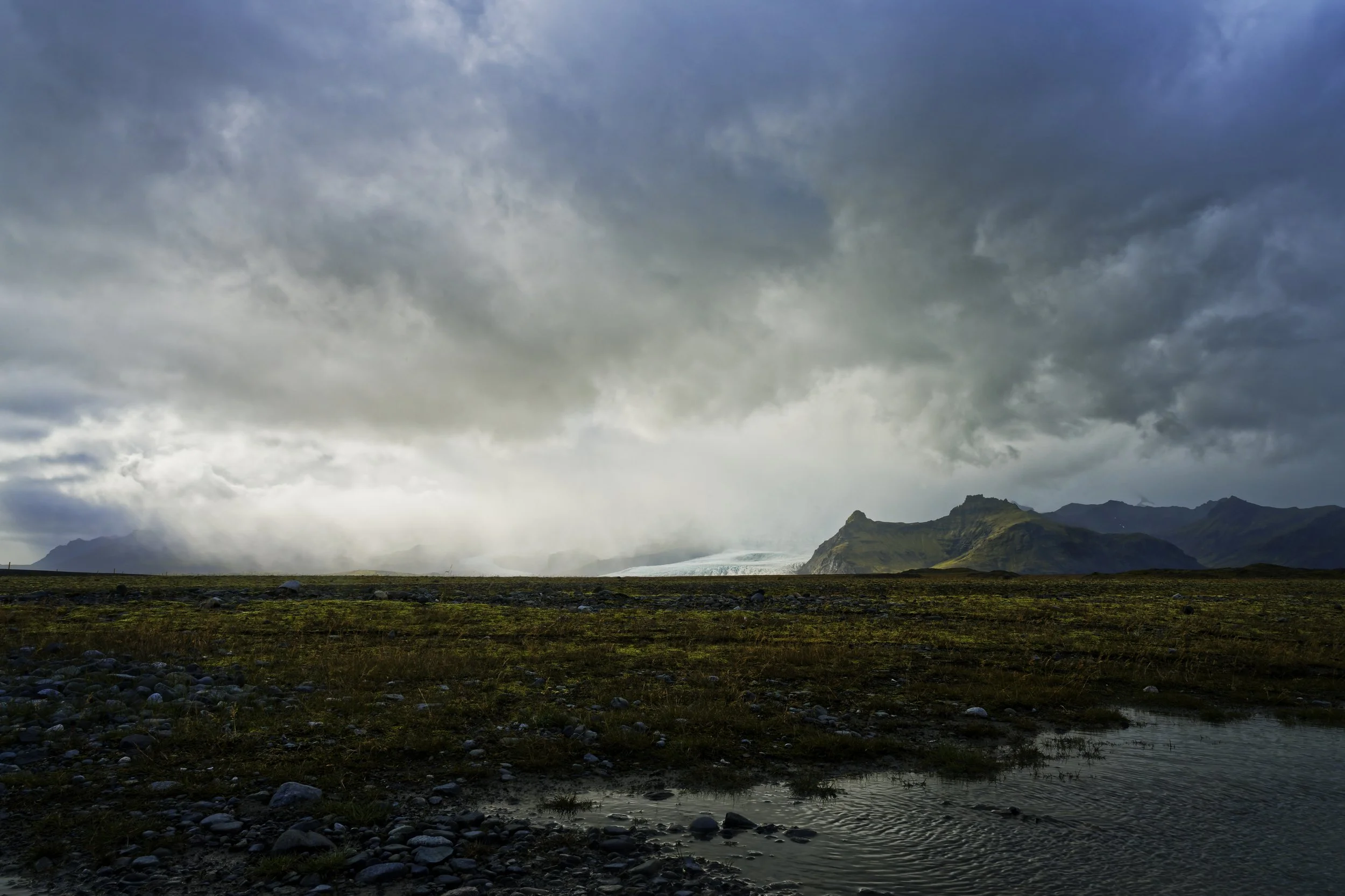 A landscape scene with dark, stormy clouds over distant mountains and a small body of water in the foreground.