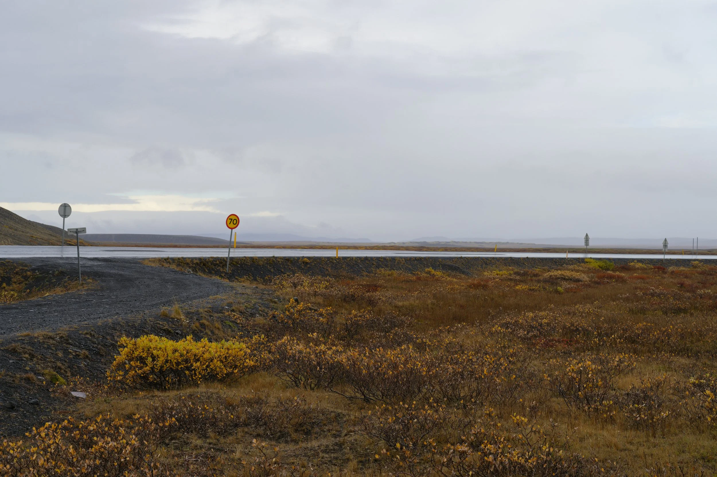 A remote highway scene with multiple road signs on the side, a speed limit of 70, and a cloudy sky over a flat, open landscape with sparse vegetation.