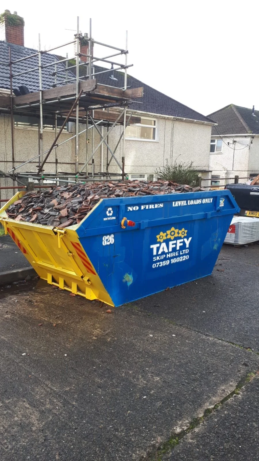 Construction dumpster filled with roof tiles and stone debris in front of a house with scaffolding for roof work.
