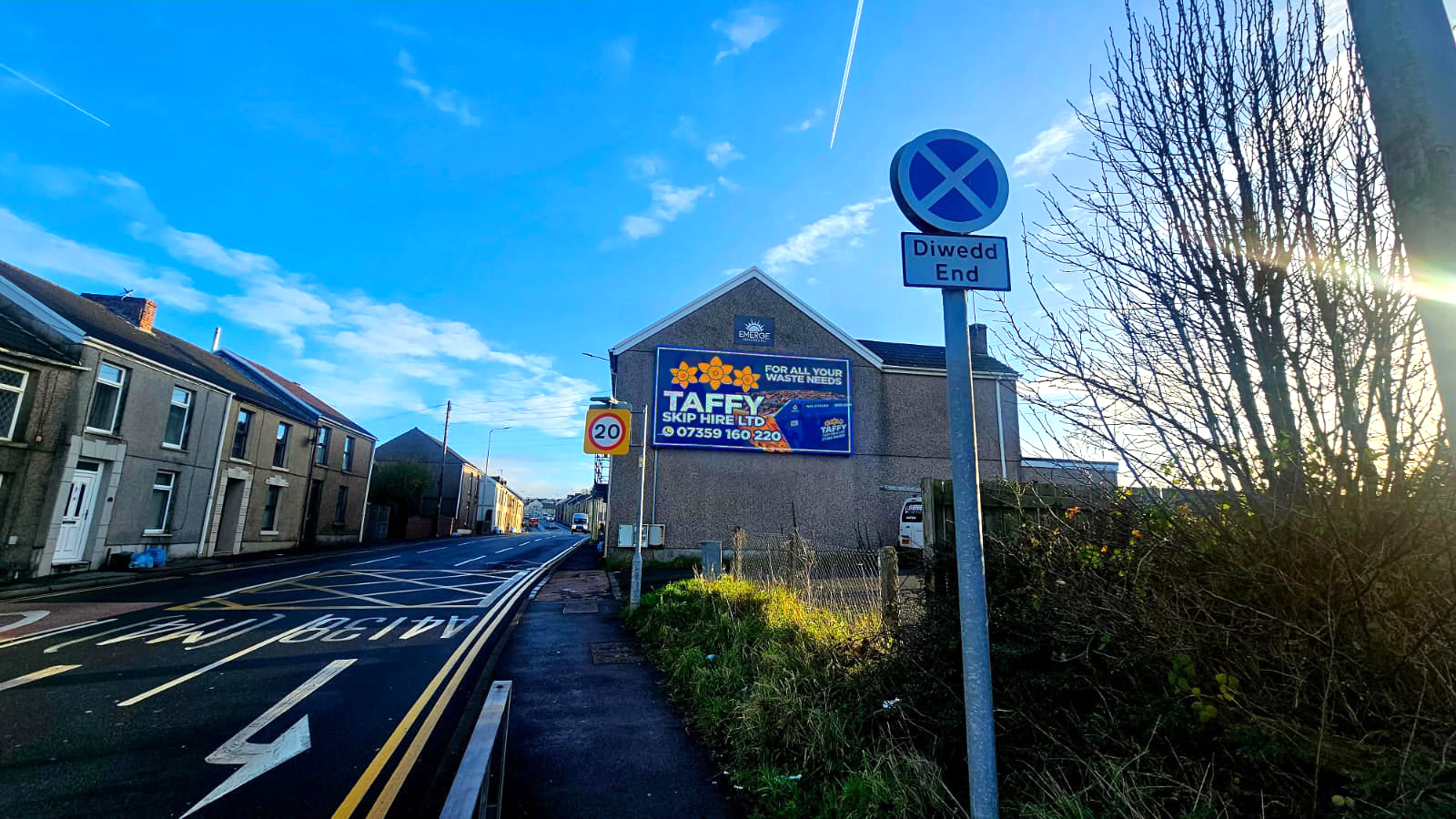 A street scene in a residential area with a no parking sign and a speed limit sign of 20 miles per hour. There is a large billboard on a building advertising a waste disposal company, and houses on the left side of the street. The sky is blue with some clouds, and the sun is shining through trees on the right.