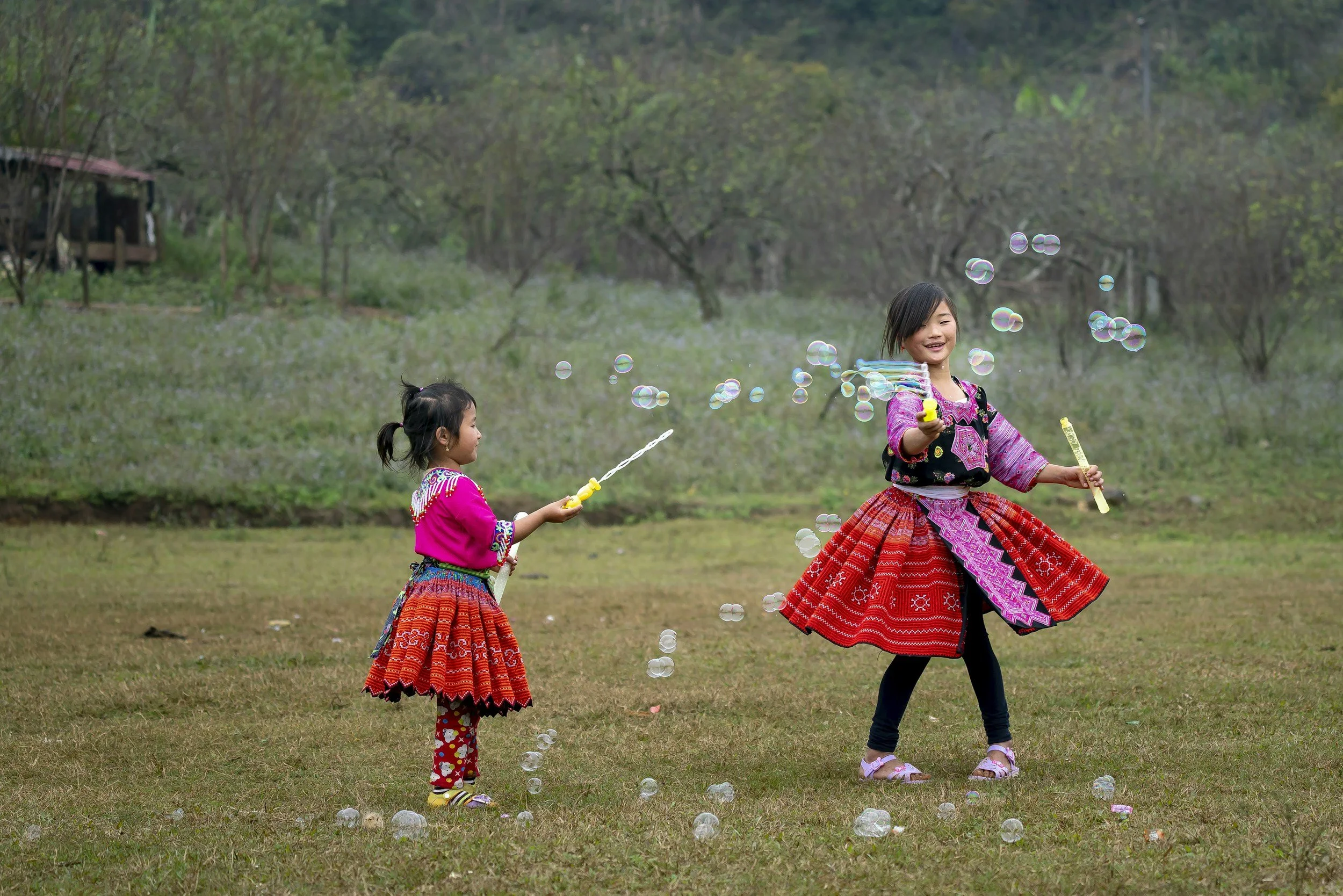 Two young girls in traditional colorful clothing playing with bubbles in an outdoor grassy area with trees in the background.