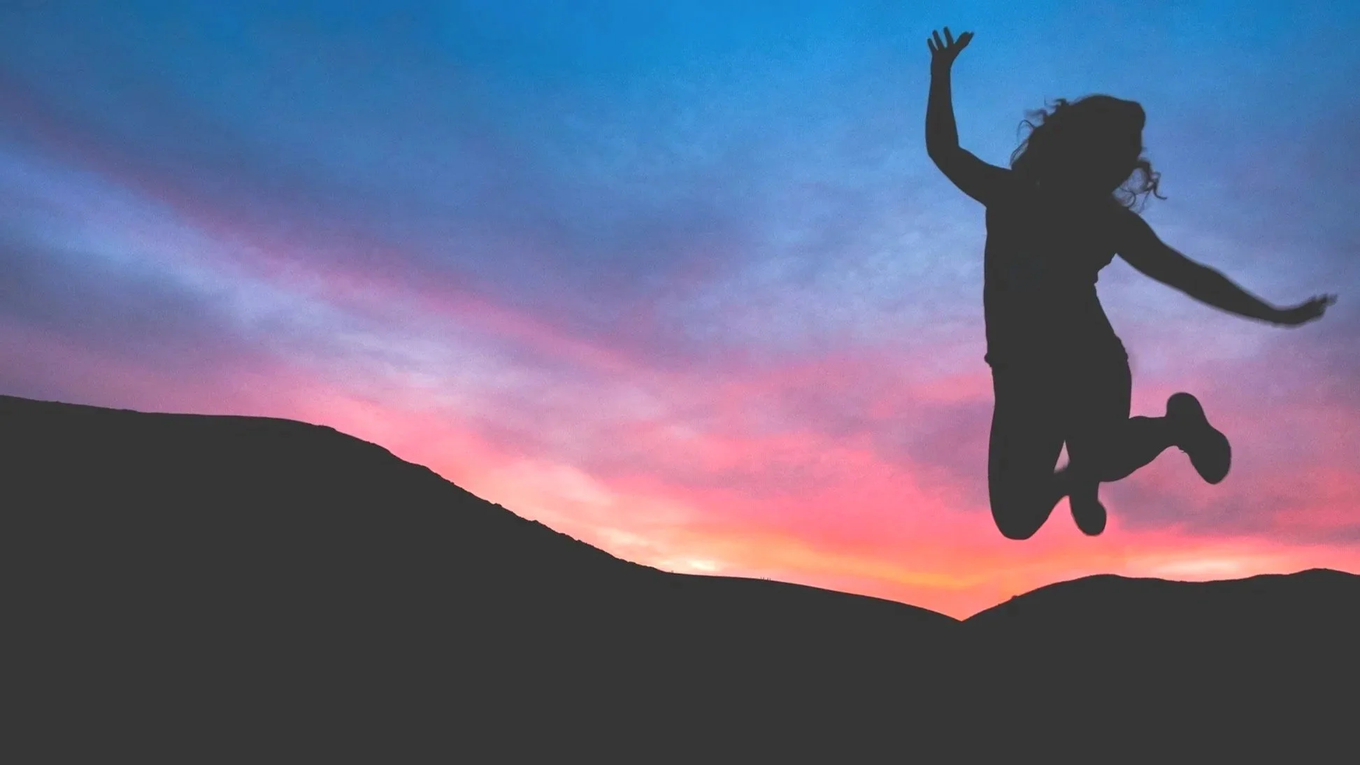 Silhouette of a girl jumping in the air against a colorful sunset sky with hills in the background.