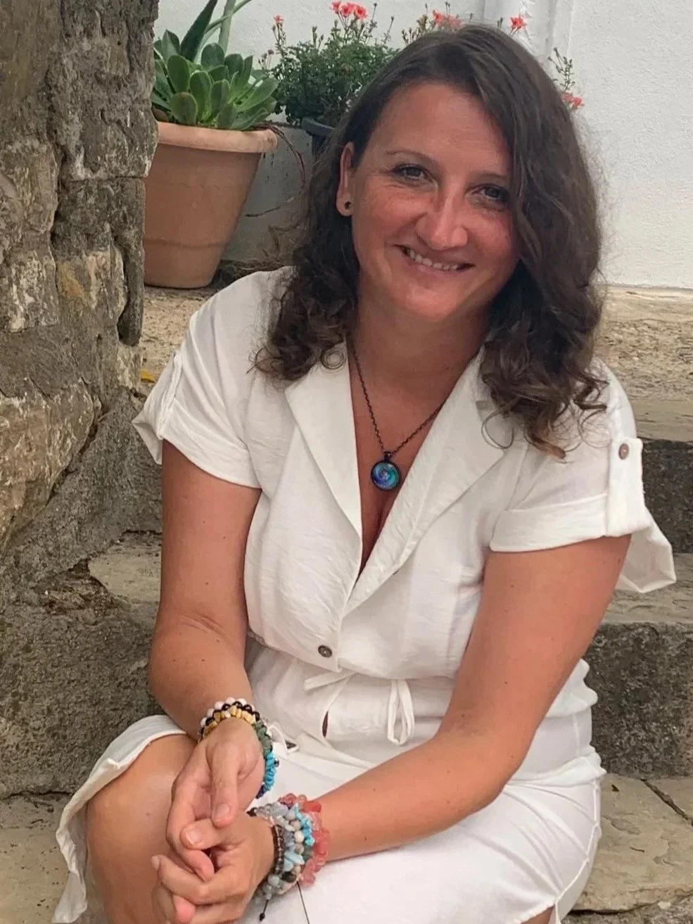 A woman with dark, curly hair, wearing a white dress with a tied waist, sitting outdoors near a stone wall with potted plants and flowers in the background.