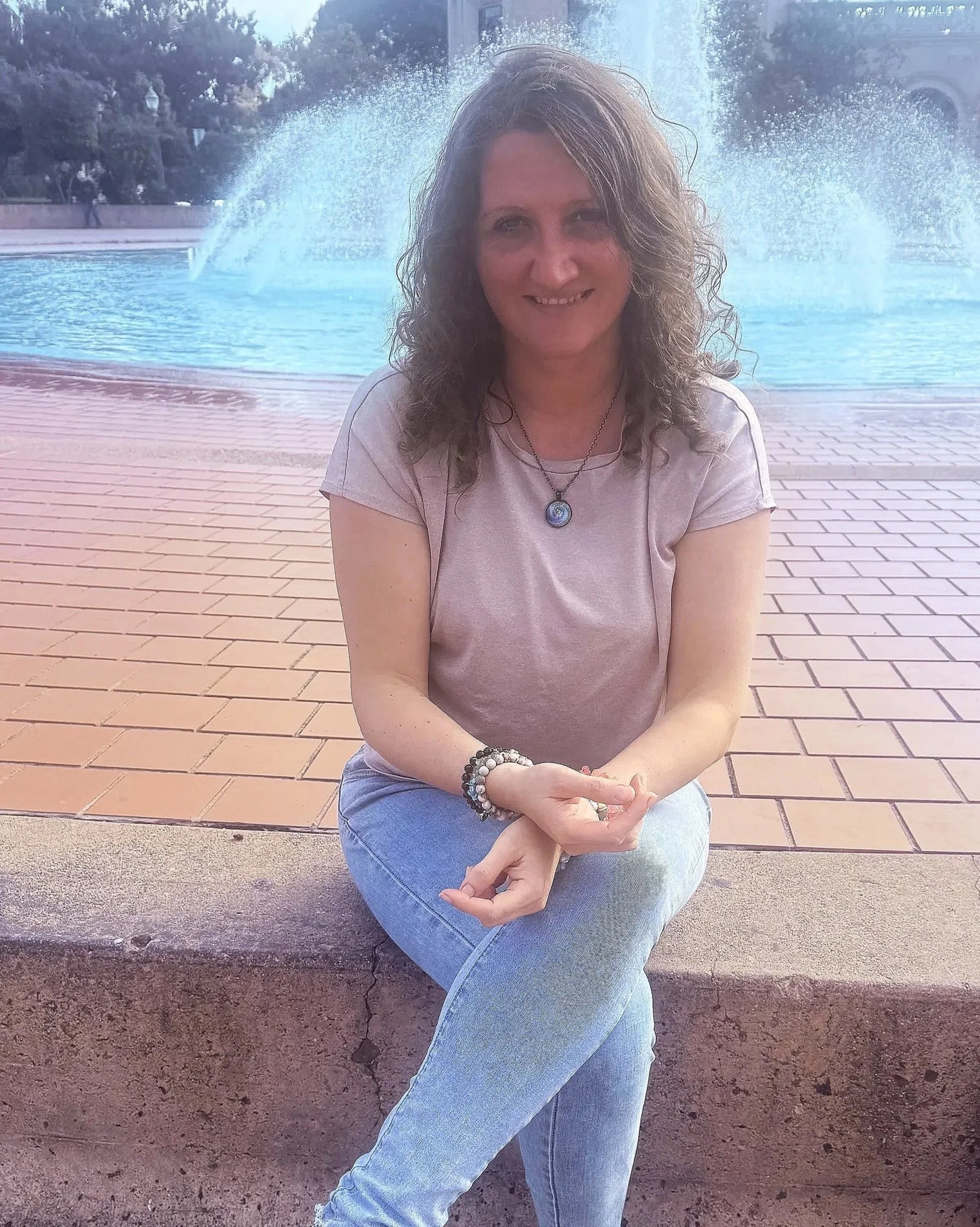 A woman with curly hair smiling, sitting cross-legged on a concrete ledge in front of a fountain with water spraying, wearing a pink t-shirt, jeans, a necklace, and bracelets.