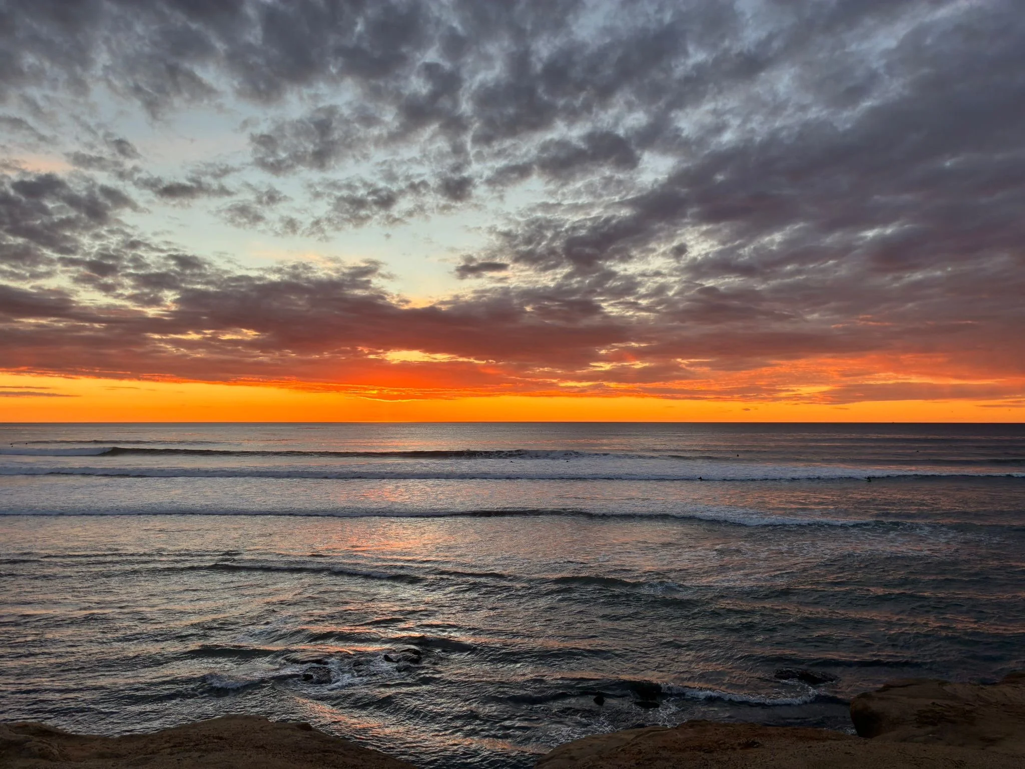 Sunset over the ocean with a partly cloudy sky, gentle waves, and rocks in the foreground.