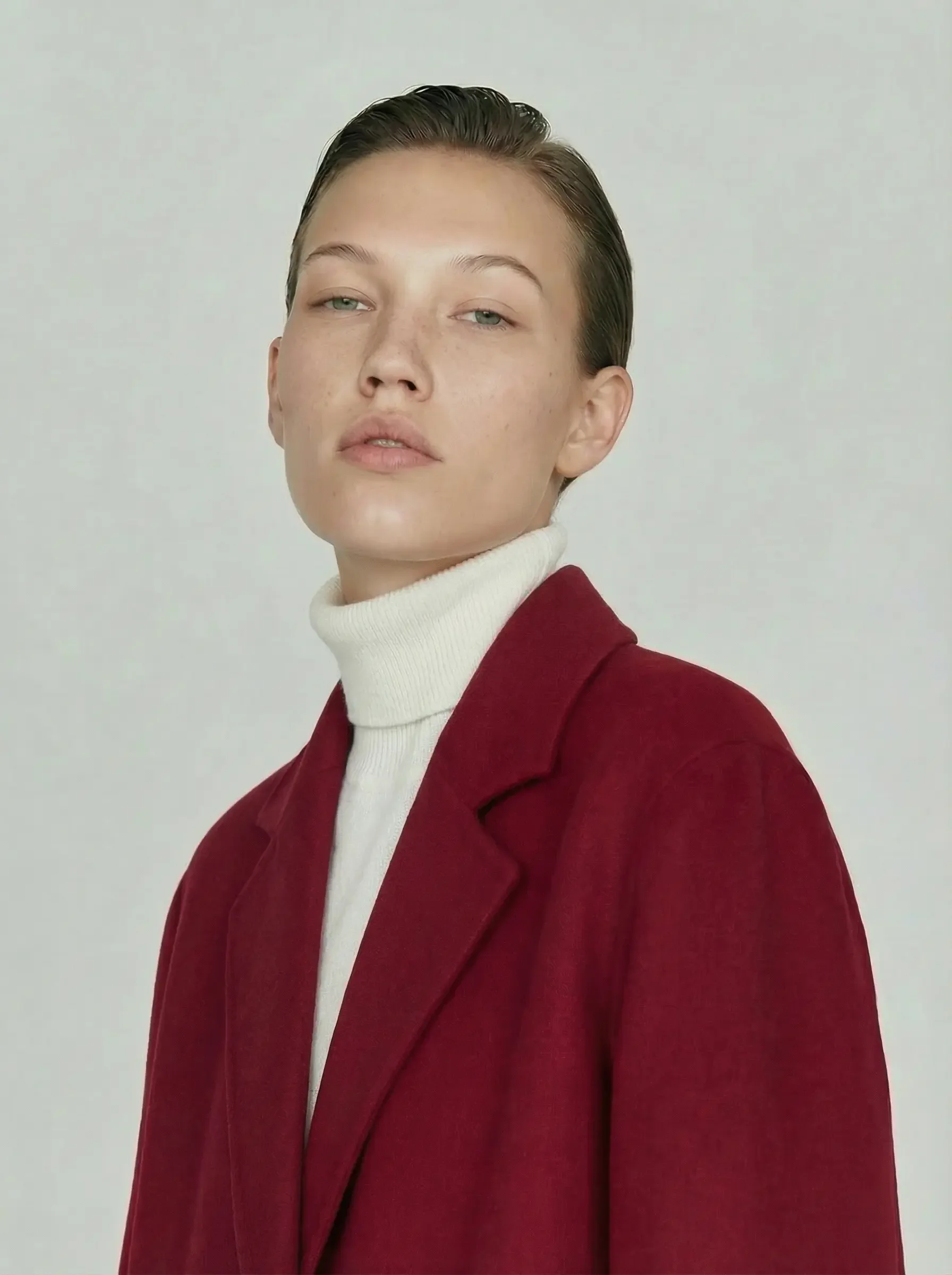 Ritratto frontale di una modella con capelli pettinati all'indietro e sguardo intenso. Indossa un maglione a collo alto color panna sotto un cappotto di lana rosso scuro dal taglio classico.