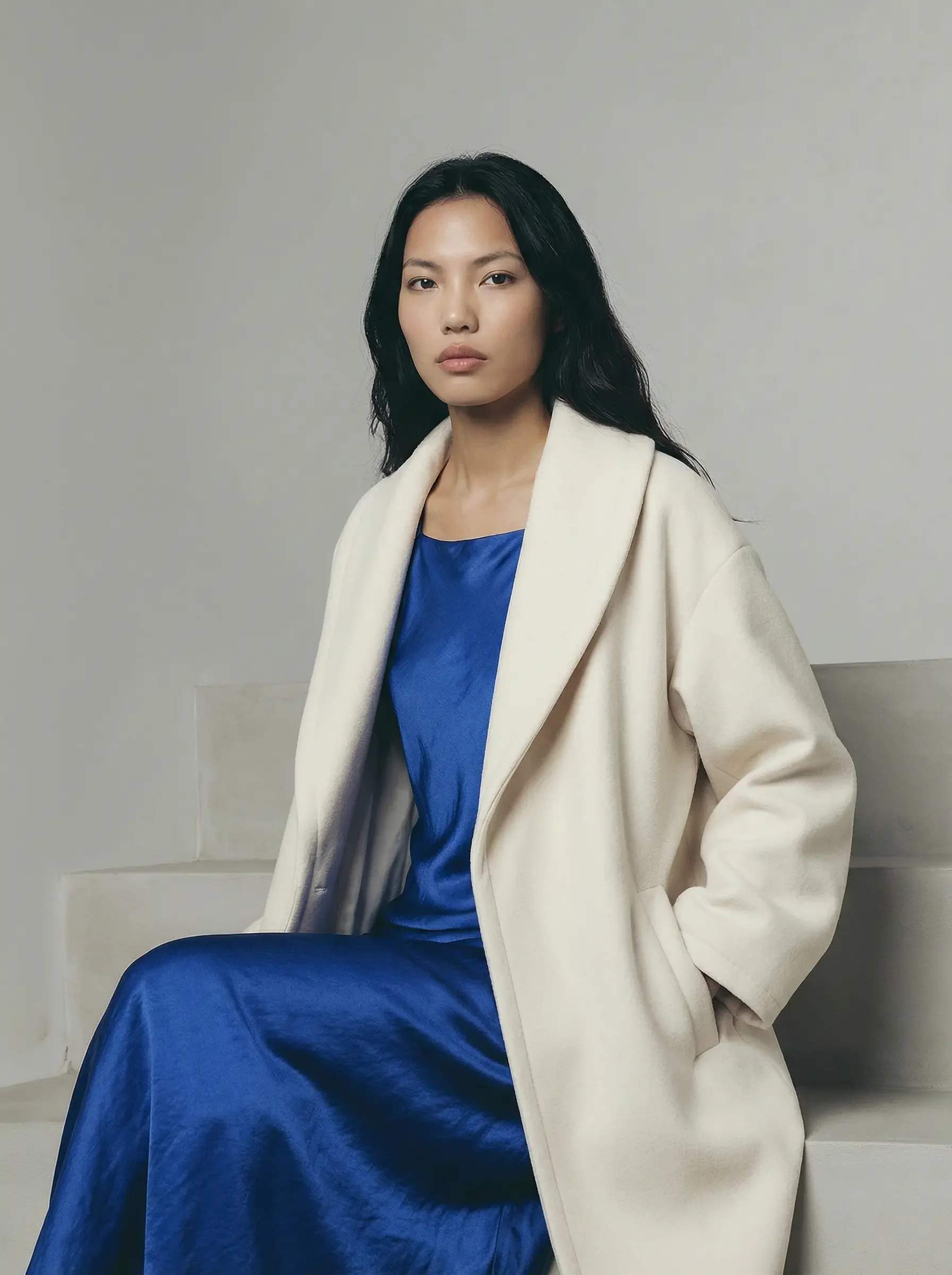 Inquadratura a mezzo busto di una donna seduta su scalini di pietra, che guarda direttamente verso l'obiettivo con un'espressione neutra. Veste un abito di seta blu elettrico e un cappotto lungo color crema.