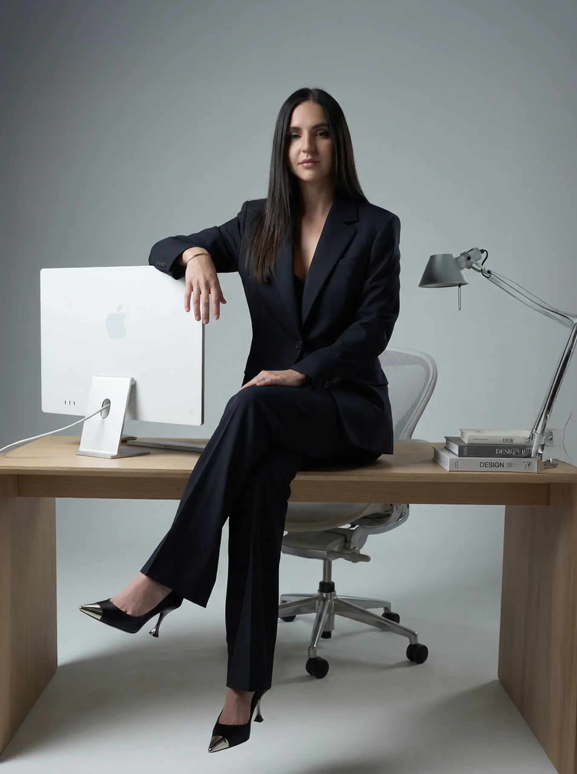 una donna seduta su un tavolo di una scrivania appoggiata con un braccio a un computer mac è vestita con un taller nero e delle scarpe con il tacco nere e la punta argento