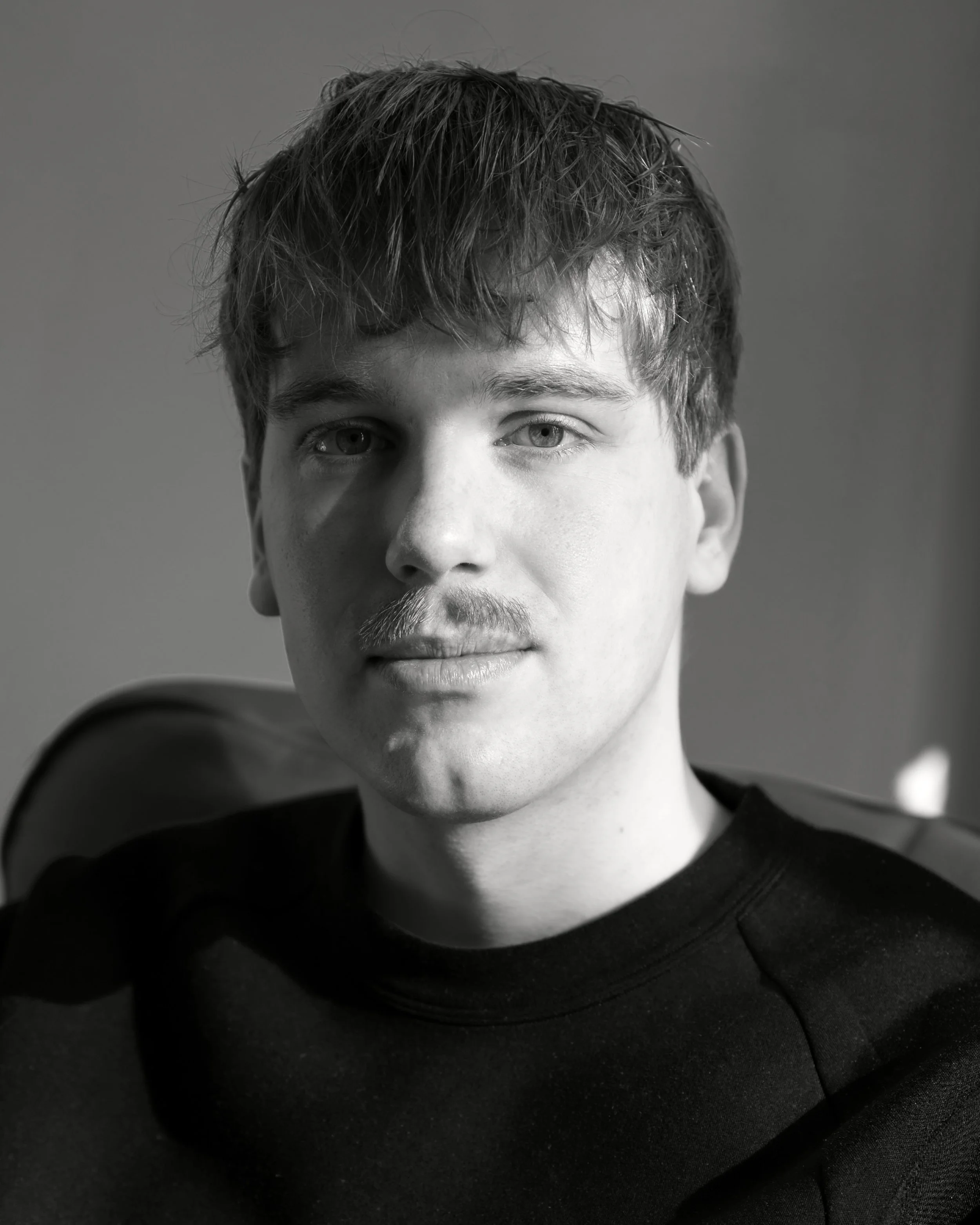 Black and white portrait of a young man with short, tousled hair, wearing a dark shirt, sitting on a chair, looking at the camera.