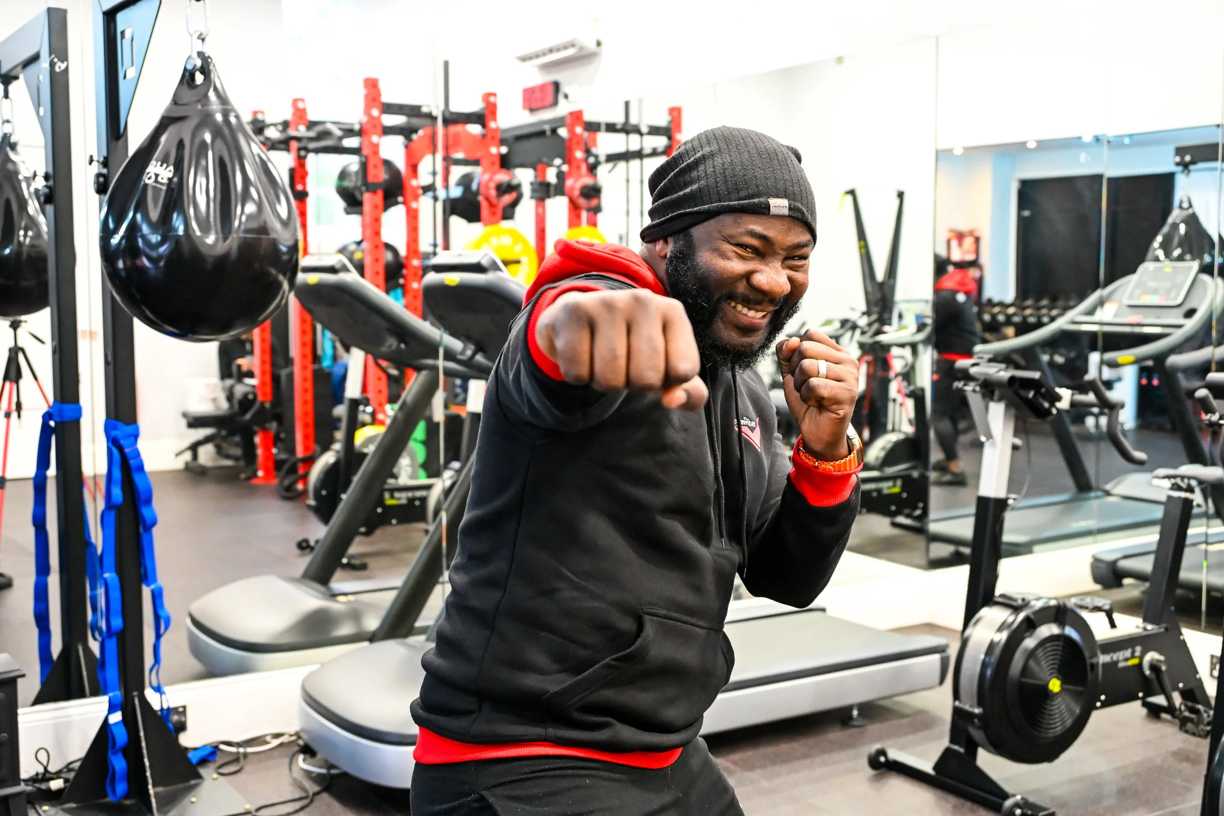 TrainRite founder, Emmanuel, wearing a black beanie and a black hoodie with red accents, posing with fists up in a fighting stance, smiling at the camera.