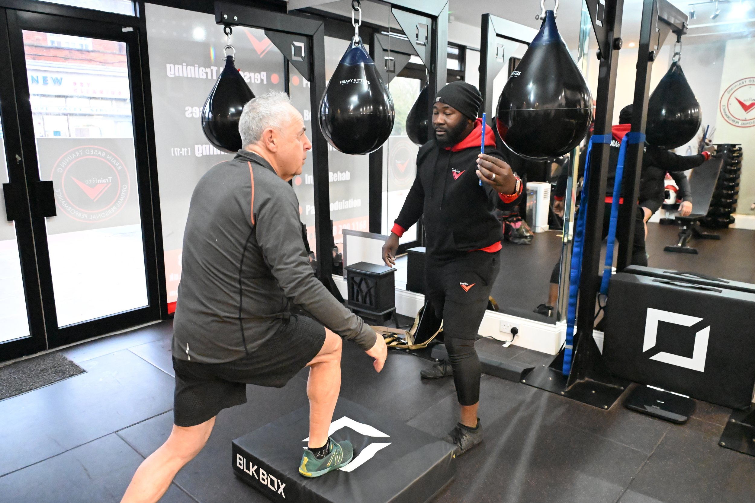 A trainer helps a man perform a step-up exercise on a platform at a gym, with punching bags hanging in the background.