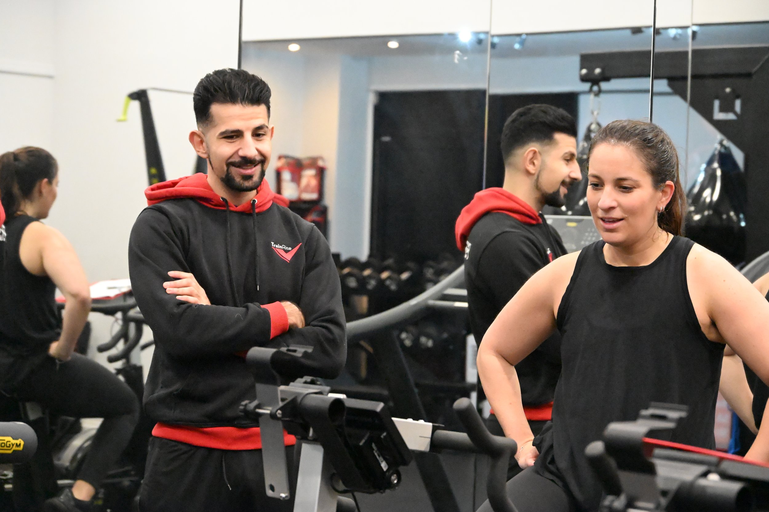 A group of people working out at a gym, including a man with a beard and dark hair wearing a black hoodie with red accents, talking to a woman with brown hair in a ponytail wearing a black athletic top, with others exercising in the background.