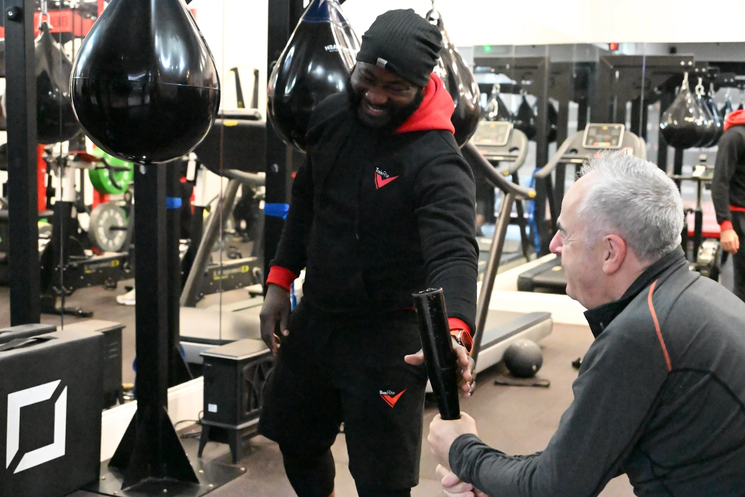 Two men smiling and having a conversation in a gym with boxing bags and workout equipment in the background.