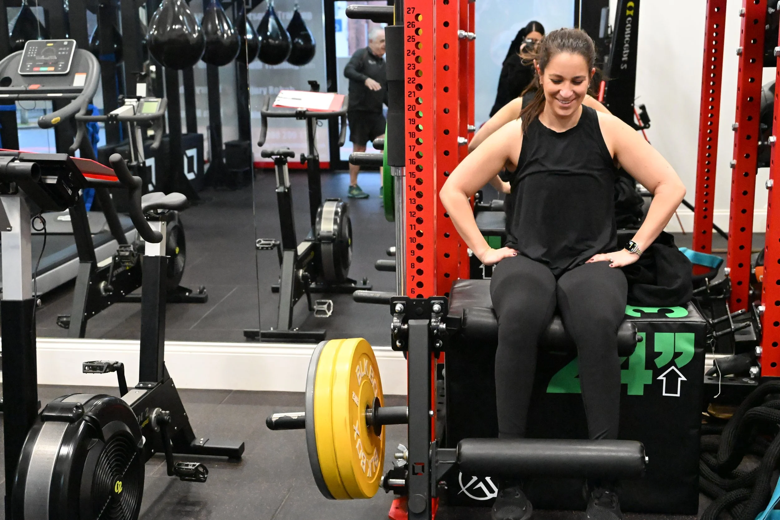 A TrainRite client sitting on a workout bench with her hands on her hips, smiling in a gym surrounded by exercise equipment and other people, with a barbell loaded with yellow and orange weights in front of her.