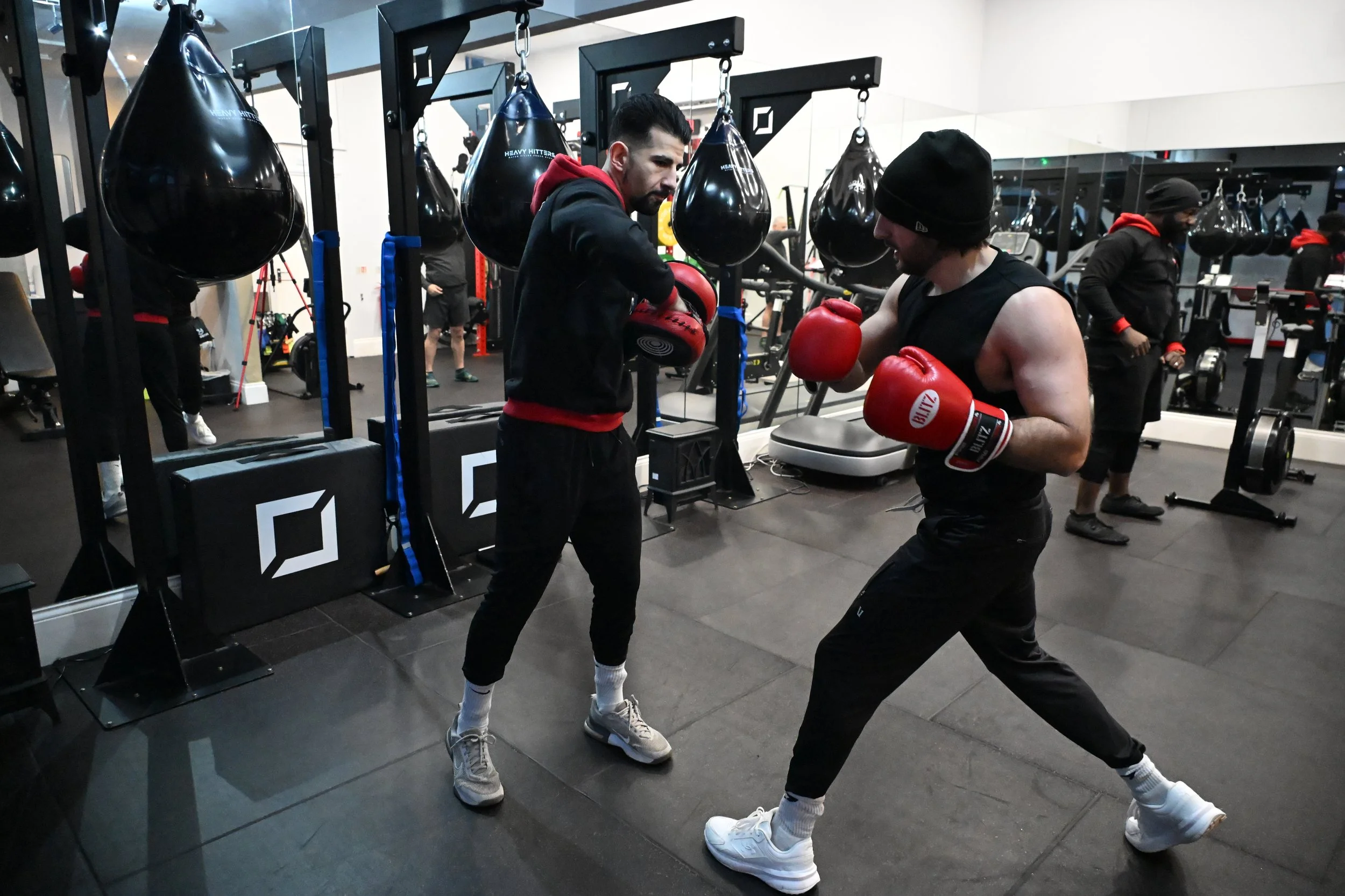 Two men boxing with red gloves in a gym, practicing with a trainer observing. Background shows punching bags and gym equipment.