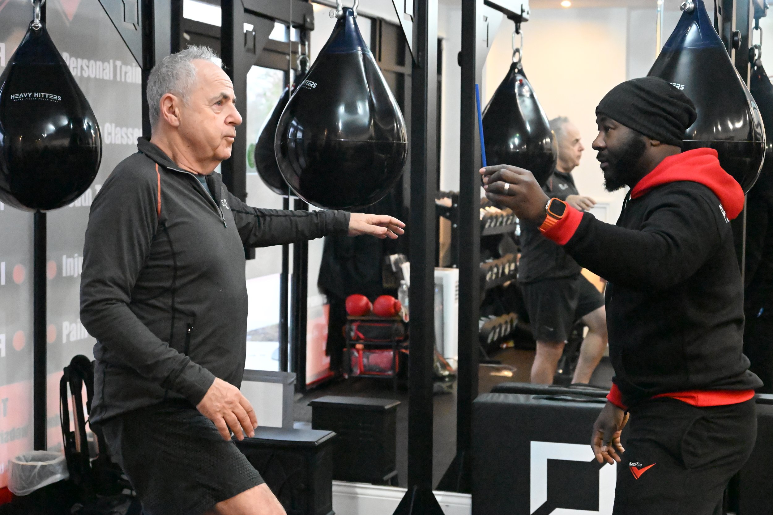A man with gray hair and dressed in black workout clothes performs a knee lift at a boxing gym, while his trainer, a Black man wearing a black beanie, black hoodie with red accents, and a smartwatch, demonstrates an exercise with a small stick among hanging punching bags.