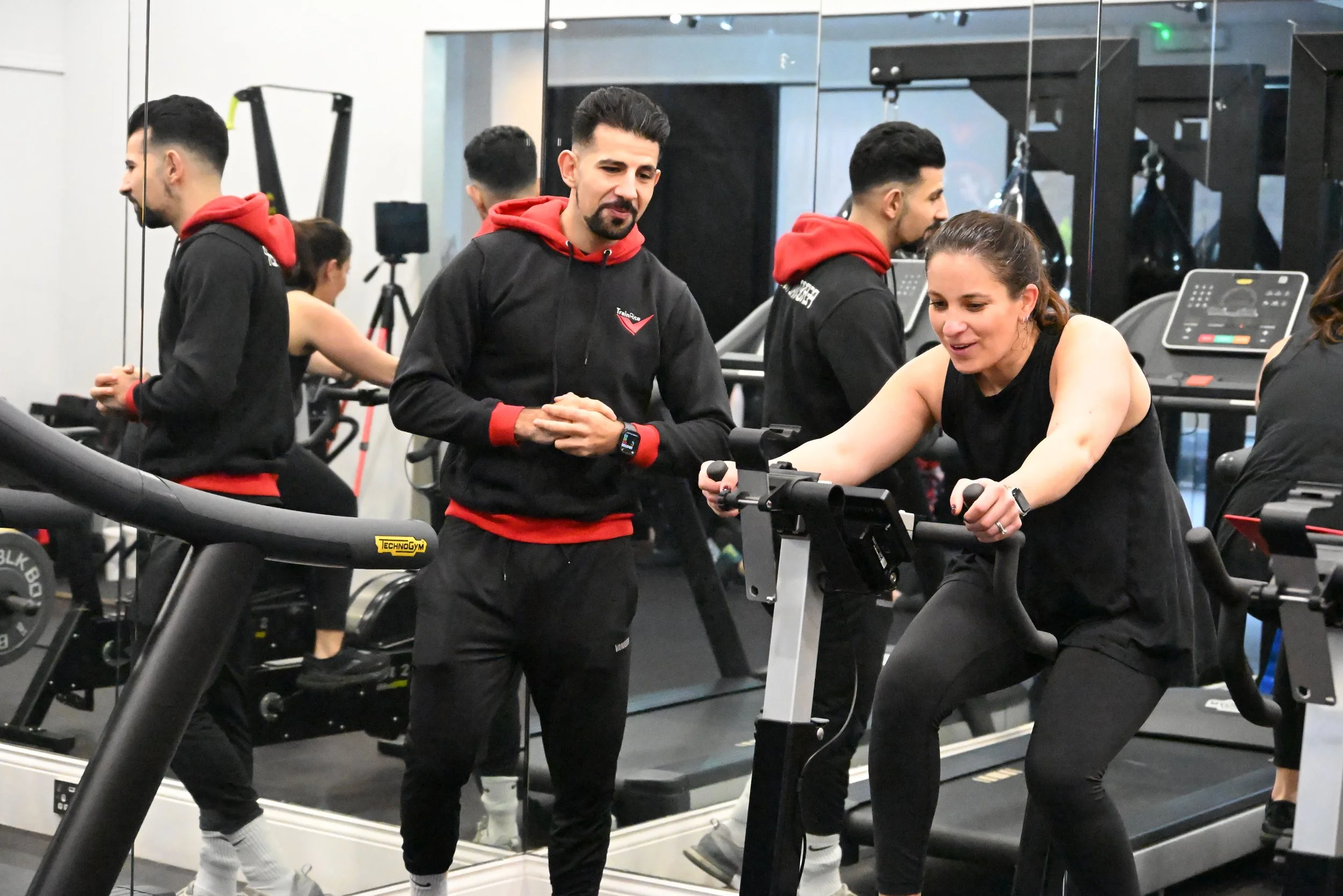 A woman working out on a stationary bike at a gym while a trainer and other individuals watch, with gym equipment and mirrors in the background.