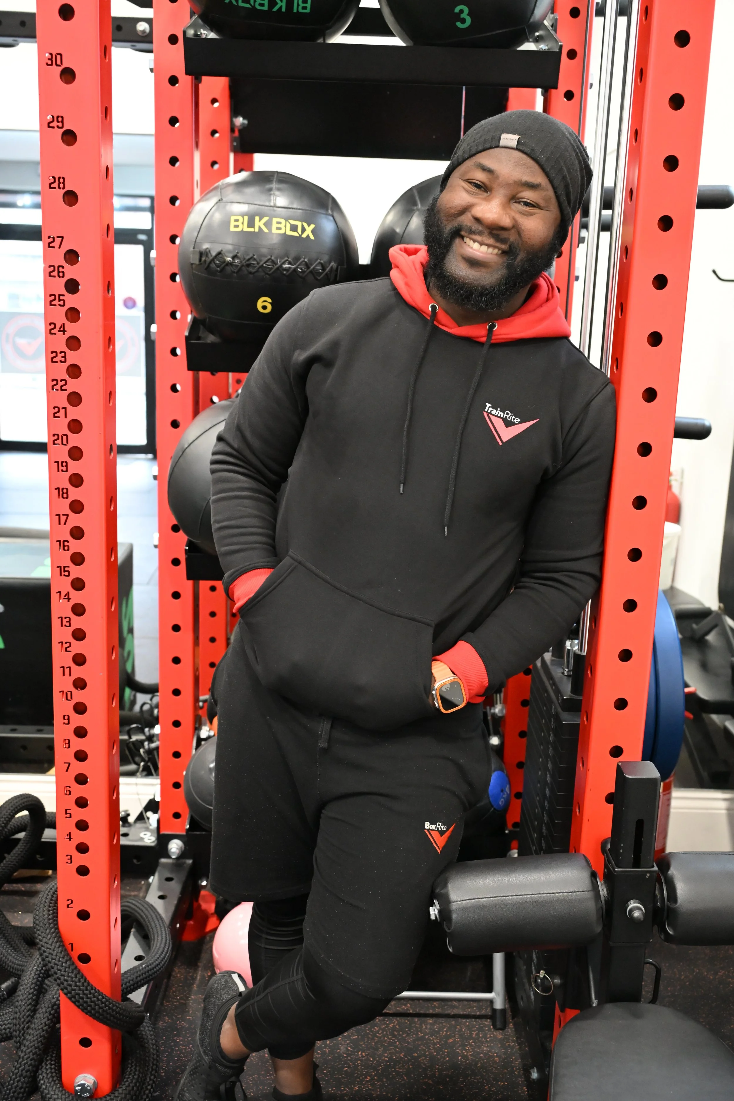 TrainRite founder and lead coach, Emmanuel, standing in a gym next to a red fitness rack with black kettlebells and medicine balls.