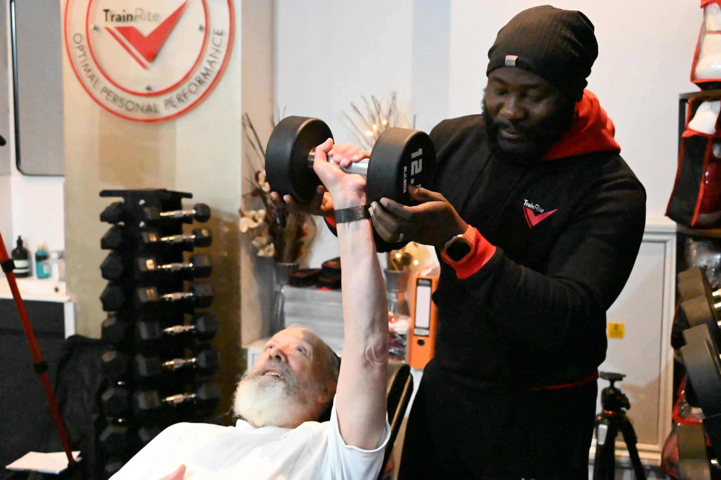 An older client lifting a dumbbell while lead coach, Emmanuel, helps him by supporting the dumbbell in a gym or fitness setting.