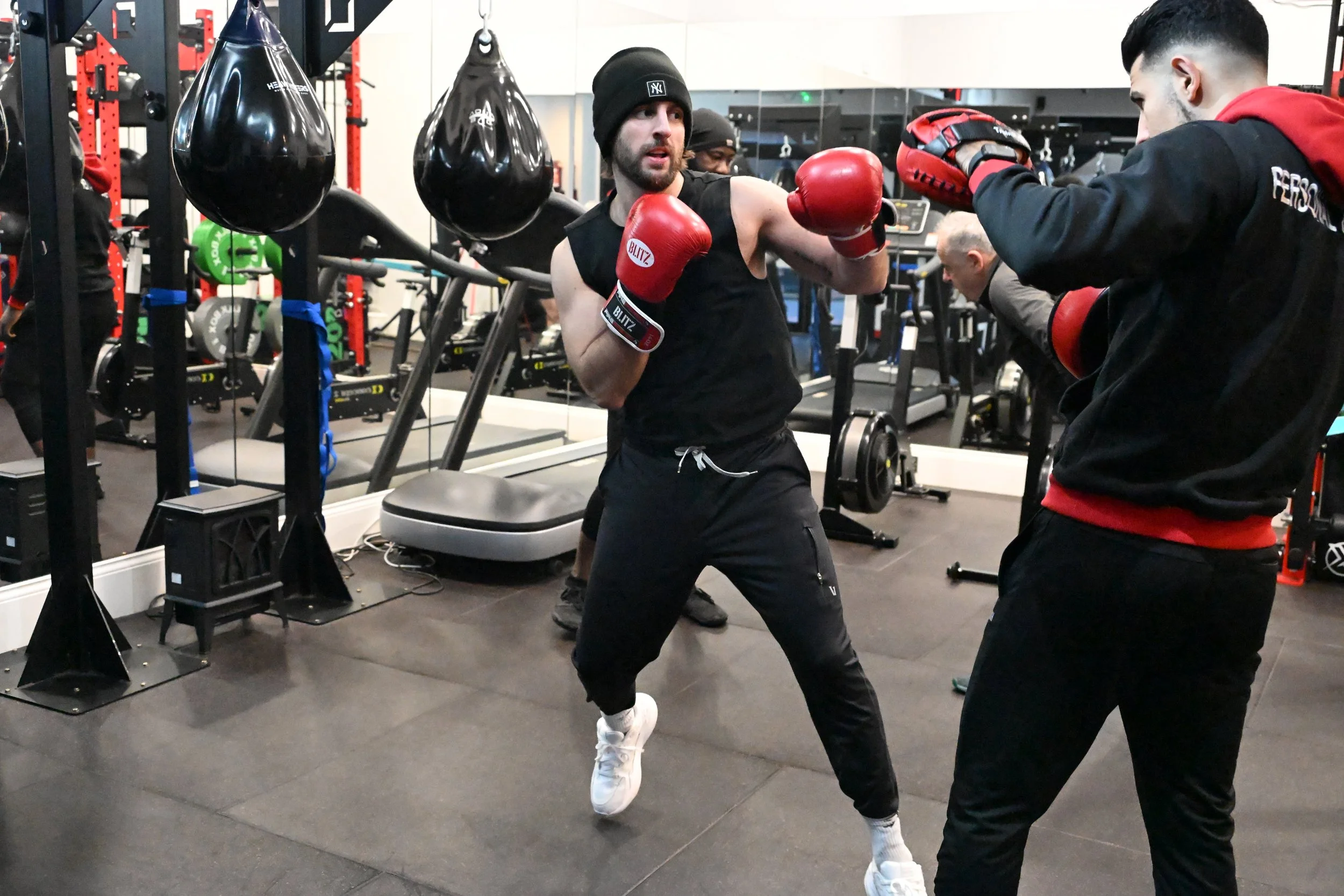 Two men boxing in a gym, practicing with gloves and focus mitts, surrounded by punching bags and gym equipment.