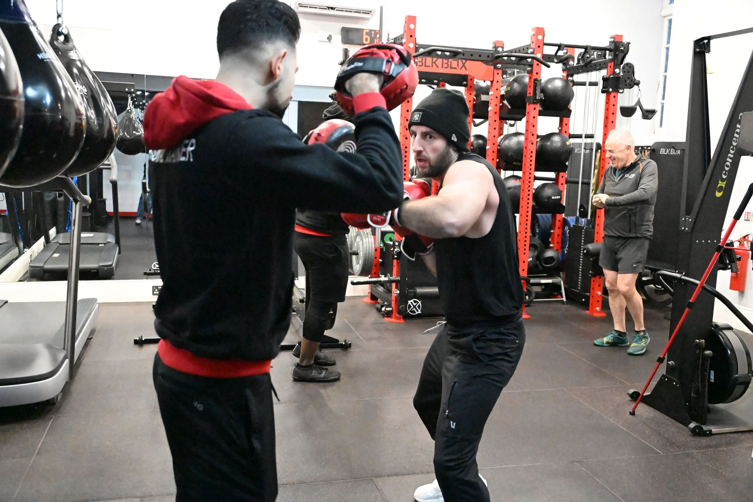 Two men practicing boxing in a gym, with gloves and headgear on, while an older man in the background watches and checks his phone.