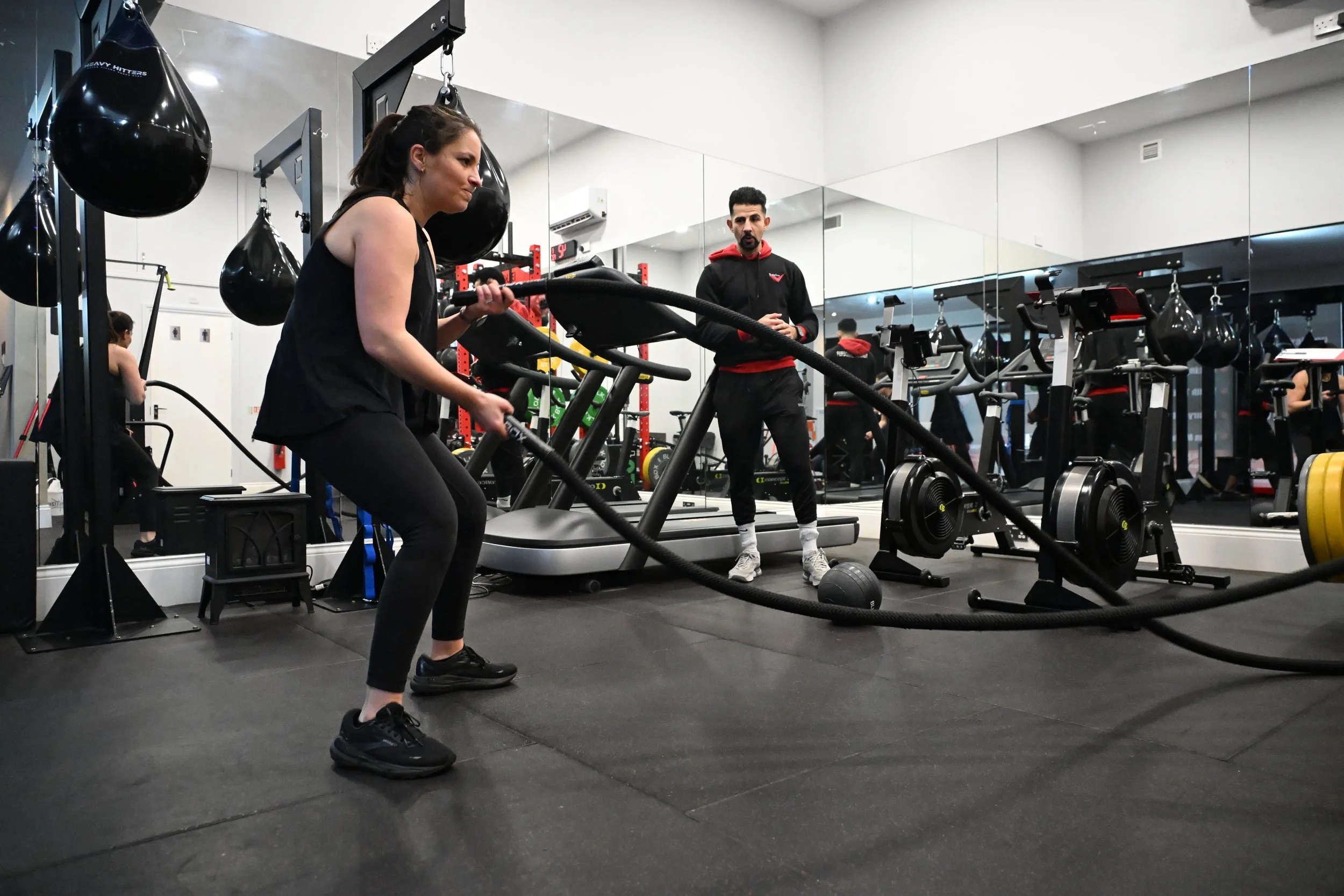 A woman exercises with battle ropes in a gym while the personal trainer observes. The gym has cardio equipment, weights, and punching bags, with mirrors on the wall.