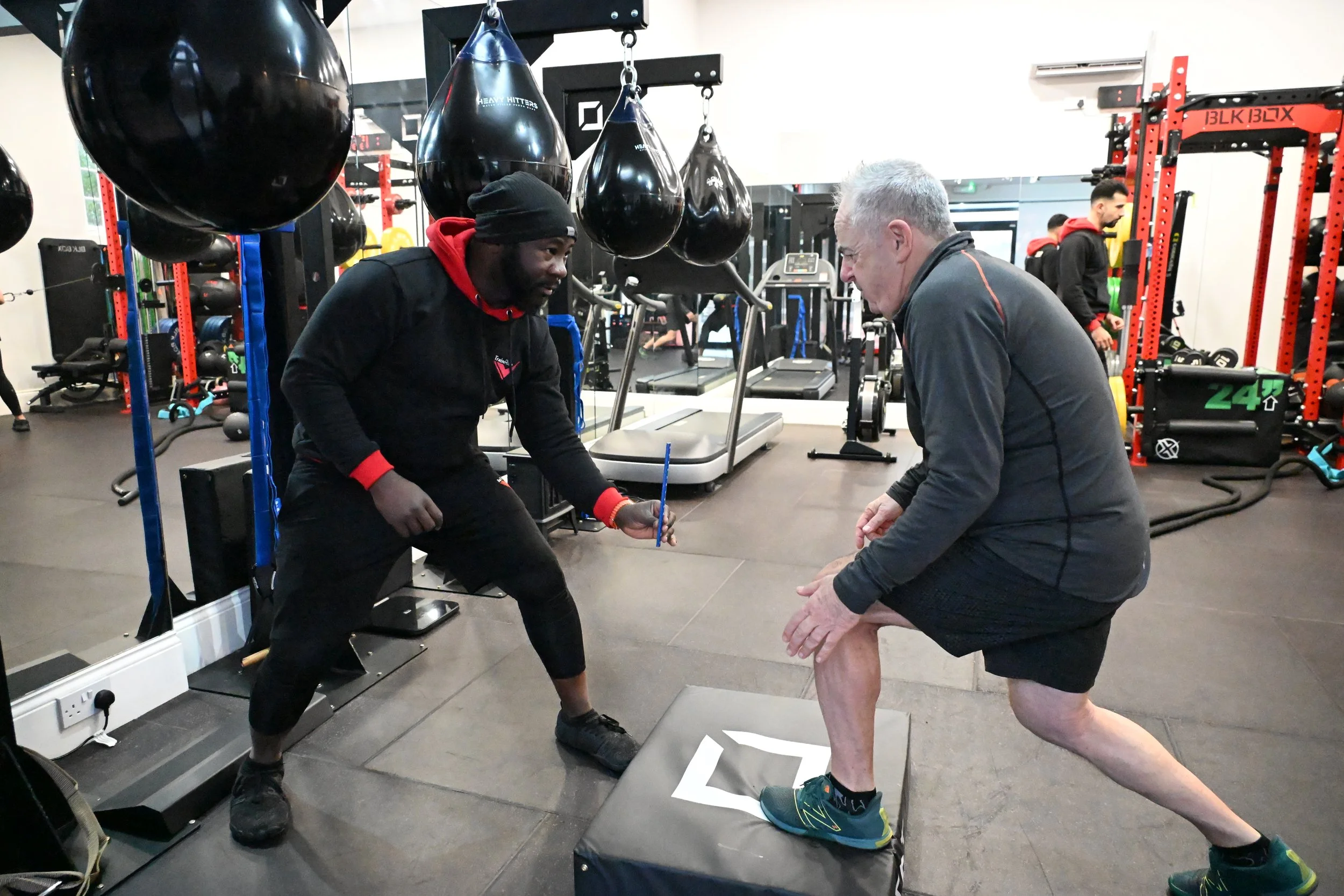 A person performing a step-up exercise on a plyometric box under the guidance of TrainRite founder, Emmanuel, in the studio, with punching bags and exercise equipment in the background.