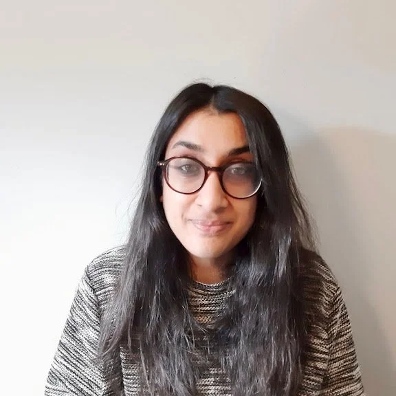 A woman with long dark hair wearing glasses and a striped sweater, smiling softly at the camera against a plain white wall.