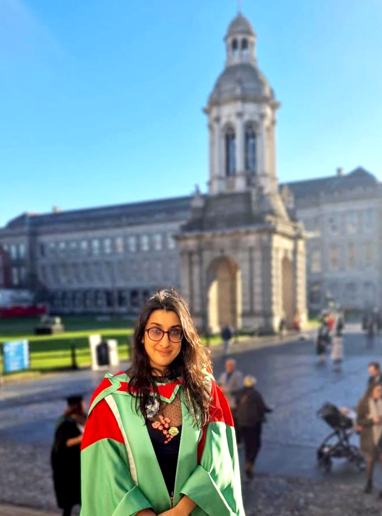 A woman with glasses and dark hair in front of a historic building, wearing a green and red graduation gown.