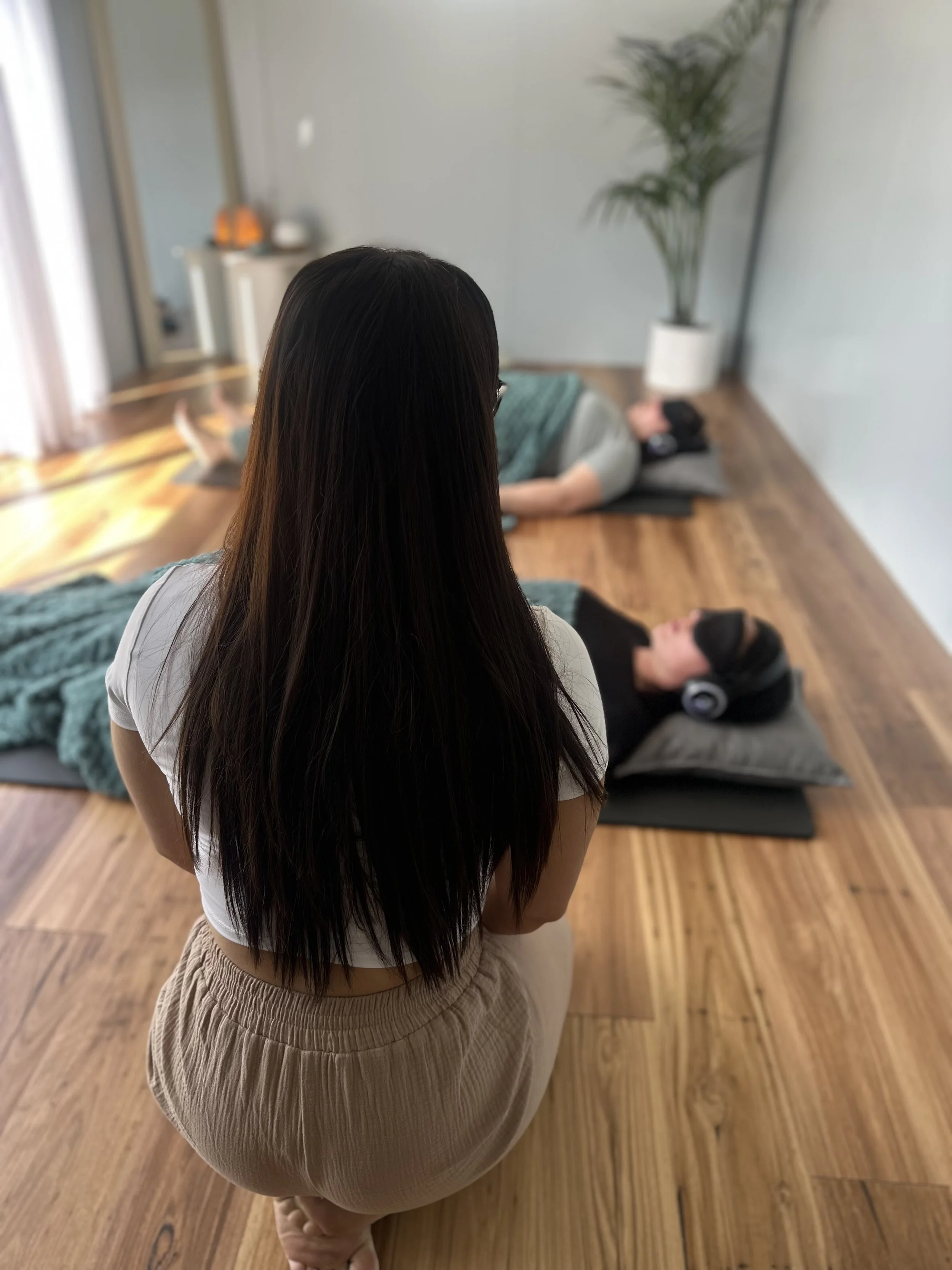 Woman with long dark hair sitting on her knees, observing two people lying on yoga mats with headphones, in a meditation or yoga class in a bright room with wooden floors, a large potted plant, and wall decor.