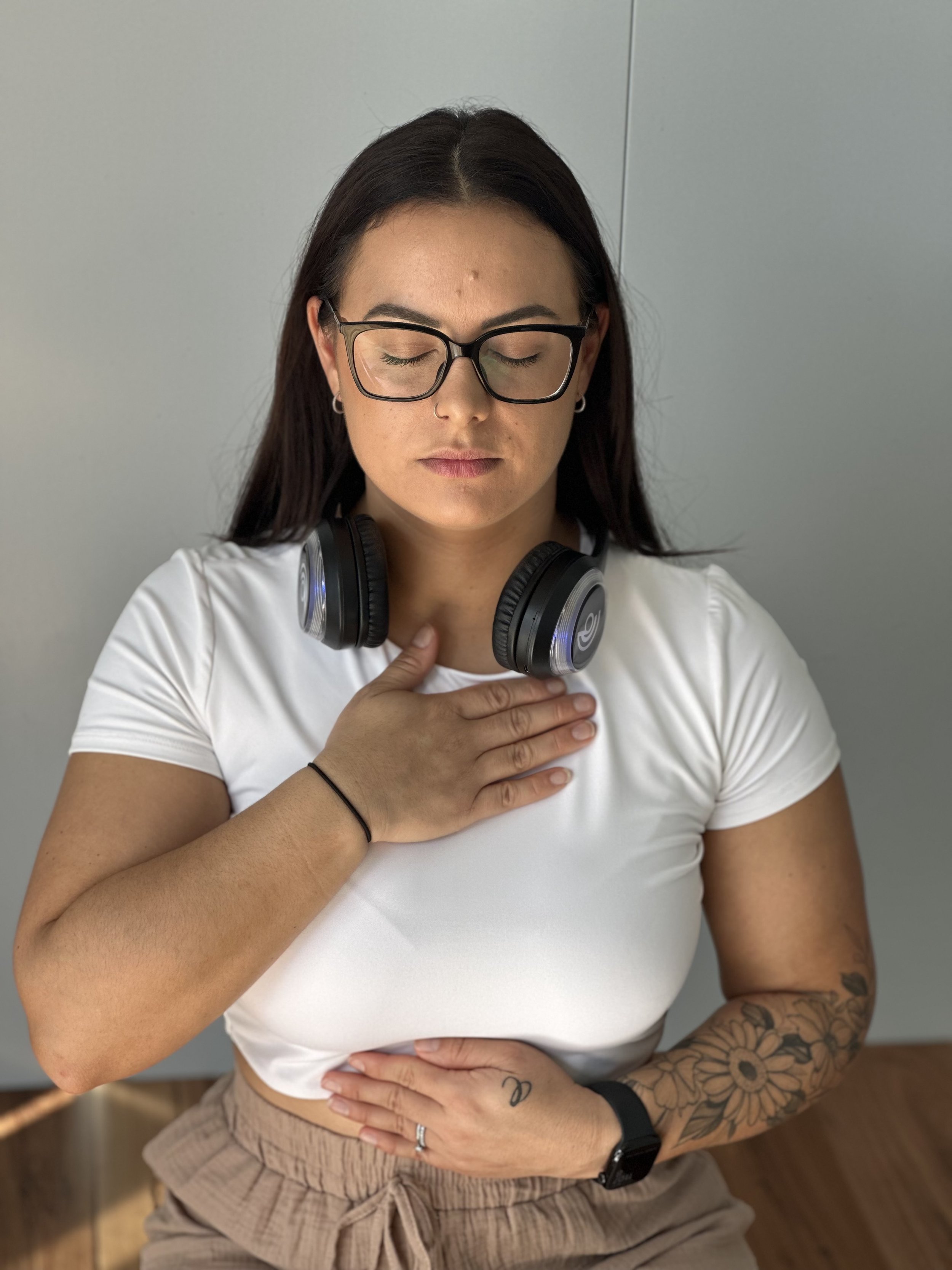 A woman with glasses, tattoos, and a black wristwatch is standing with her eyes closed and one hand on her chest, wearing a white crop top and beige shorts, with headphones around her neck.
