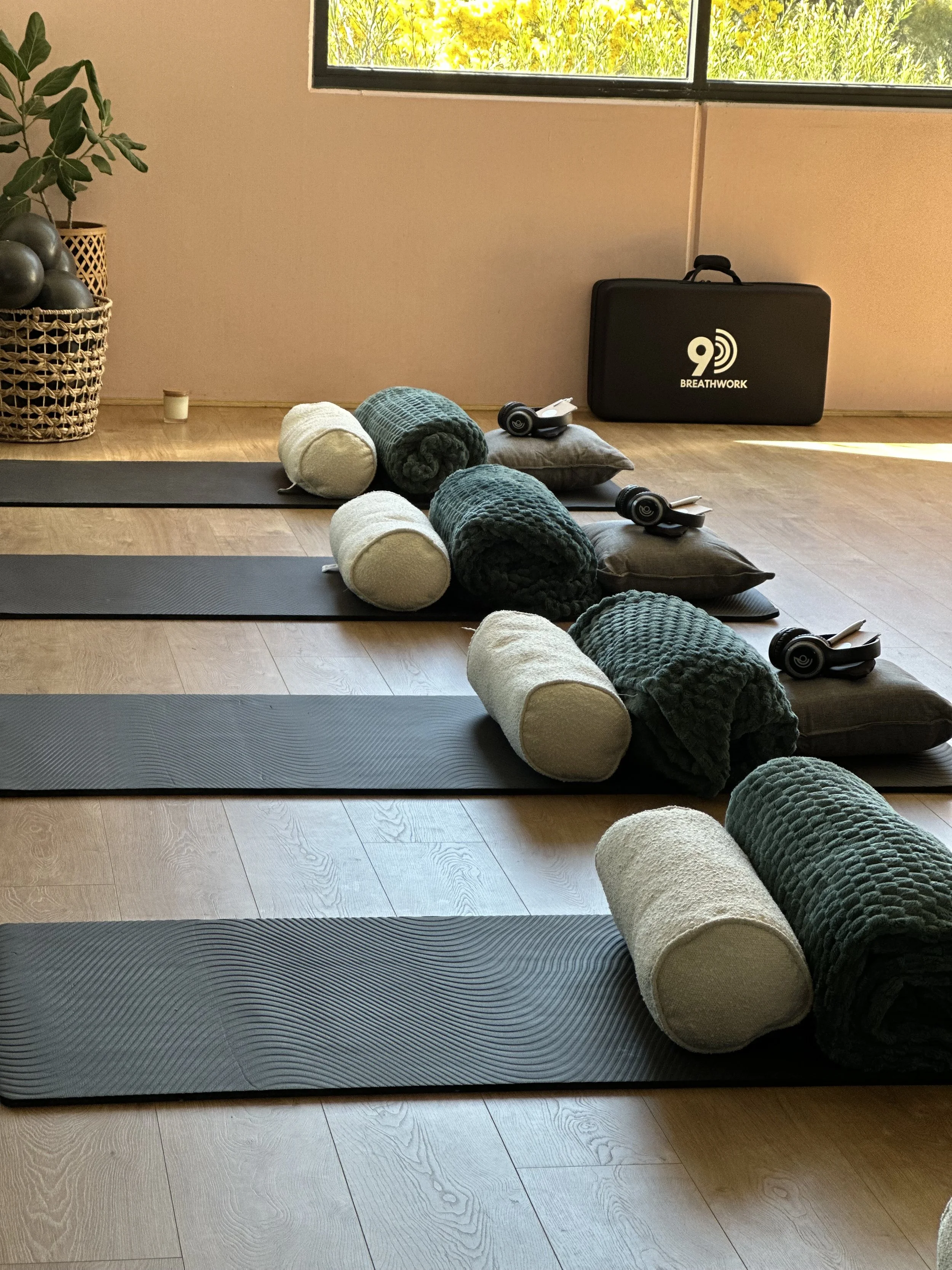 Four yoga mats with rolled towels, yoga blocks, and headphones in a yoga studio with a window and a Breathwork carrying case.